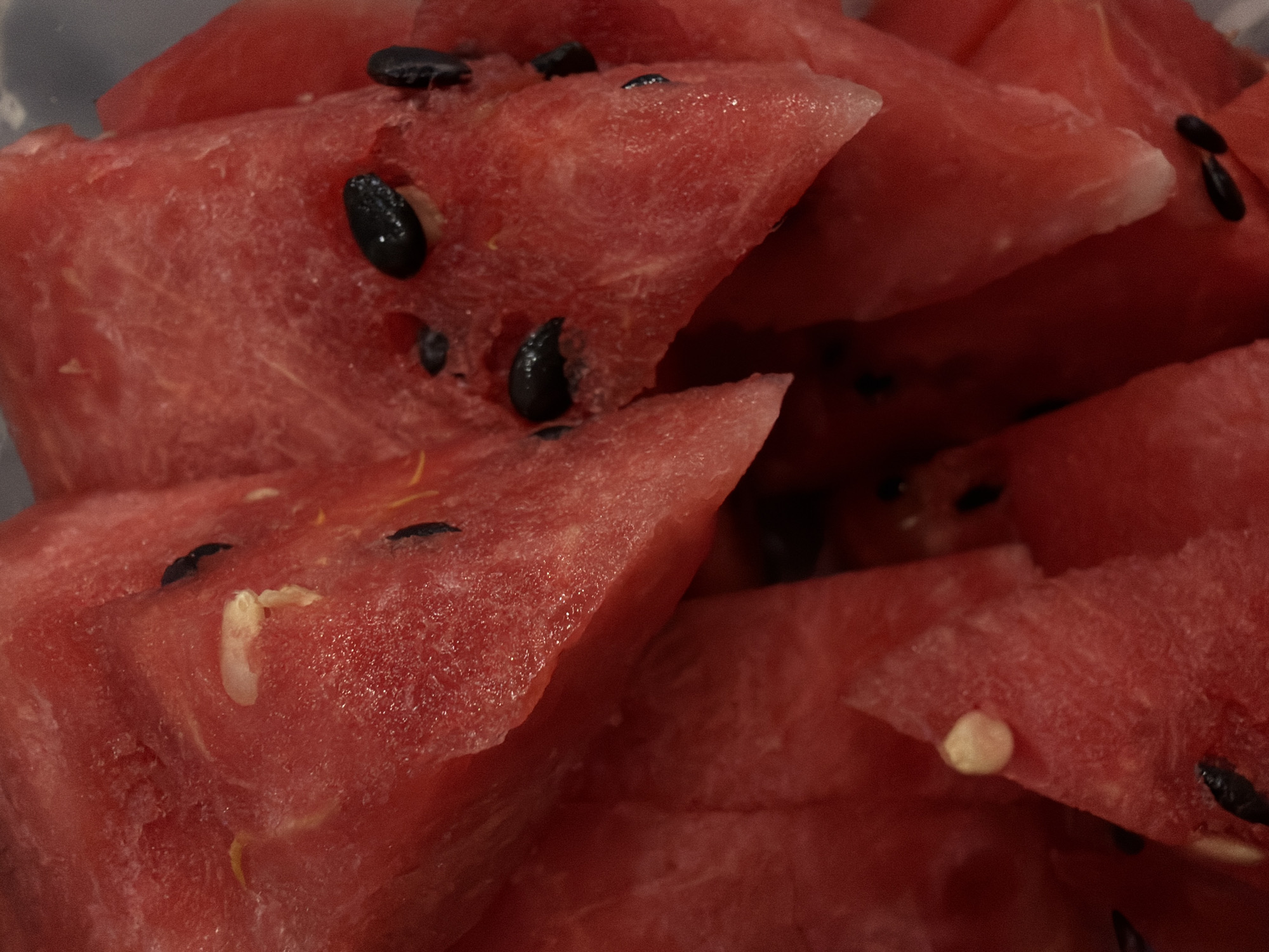The image is a close-up, overhead view of sliced watermelon. The watermelon flesh is a vibrant red, with visible white seeds scattered throughout. Some slices are triangular, while others are irregular. The texture of the watermelon appears juicy and slightly fibrous. The lighting is artificial, creating subtle highlights and shadows on the fruit. The scene is devoid of any identifiable environmental context, people, or actions, and therefore cannot be placed in Sofia, Bulgaria, or any other specific location.