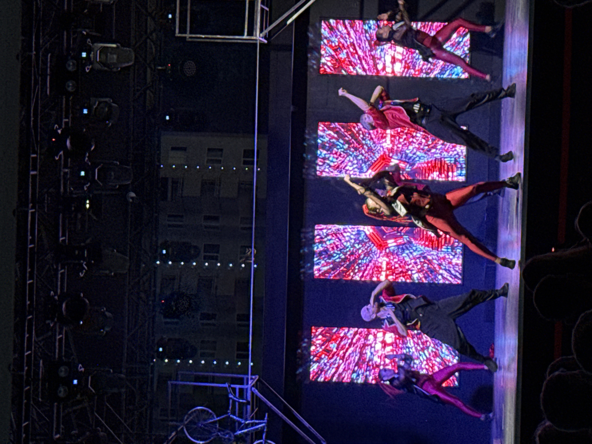This is a photograph of a stage performance in Aksu, Turkey. Six dancers, dressed in vibrant, stylized costumes of red, black, and metallic pink, are actively engaged in a choreographed routine. The stage is illuminated by a large, abstract digital backdrop displaying a psychedelic pattern of intersecting lines in pink, red, and blue. Stage lighting equipment is visible overhead. In the background, the dark silhouettes of buildings suggest the performance is taking place outdoors at night, possibly at an entertainment venue. The audience is only visible as a dark mass of heads in the foreground, indicating a live event.