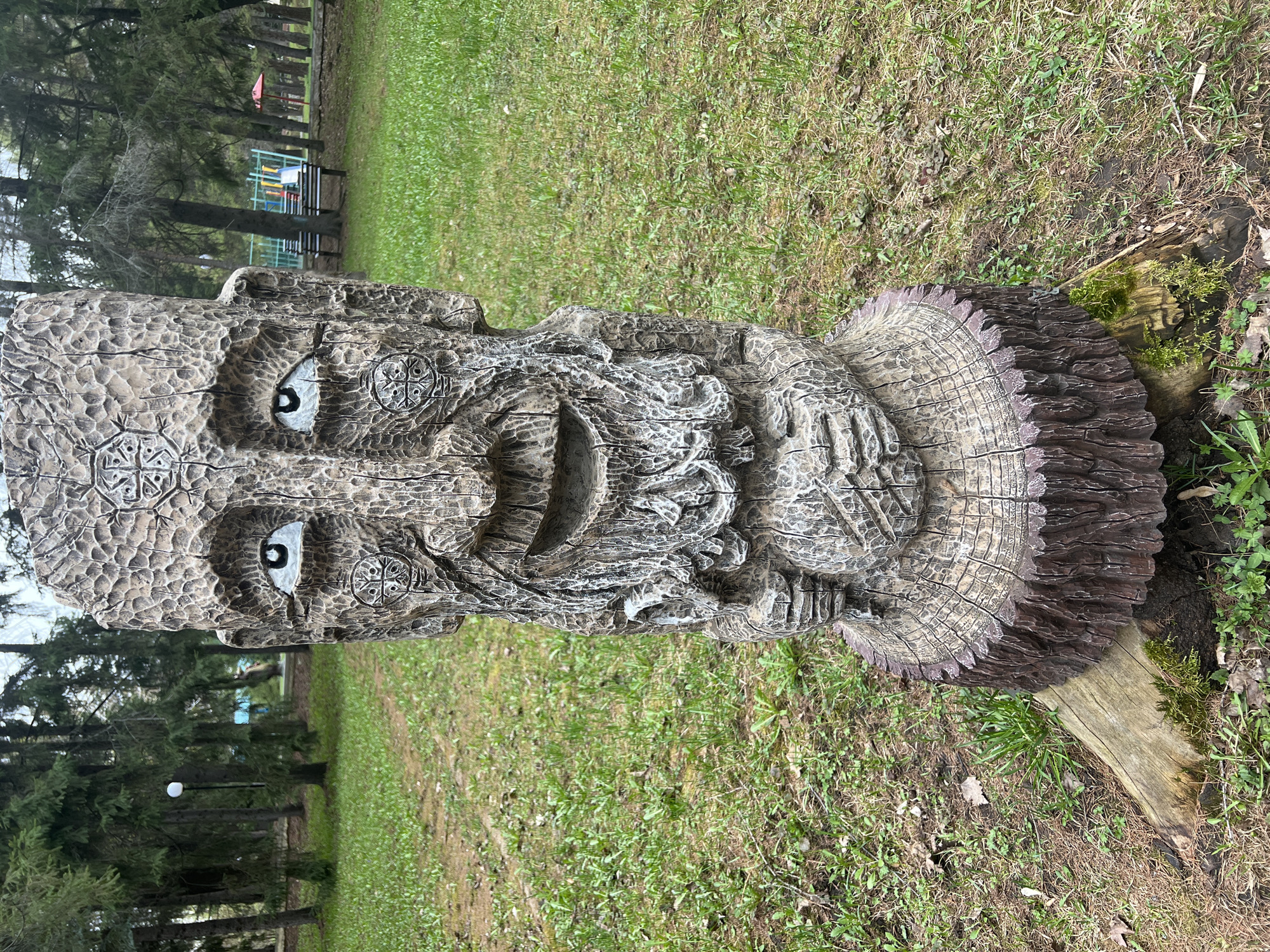 The image shows a large wooden sculpture of a man's head, likely carved from a tree trunk. The face has stylized features, including large eyes with round pupils, a smiling mouth, and a beard. Decorative symbols are carved around the eyes. The sculpture is placed on a grassy area, potentially in a park. Behind the sculpture, there are trees and possibly some playground equipment in the distance. The image is taken in Sanatoriya Yumatovo Imeni 15-Letiya Bassr, Russia. The sculpture appears to be set on a wooden support and the stump of the original tree, indicating it's a stationary installation. Moss can be seen growing near the base of the sculpture.