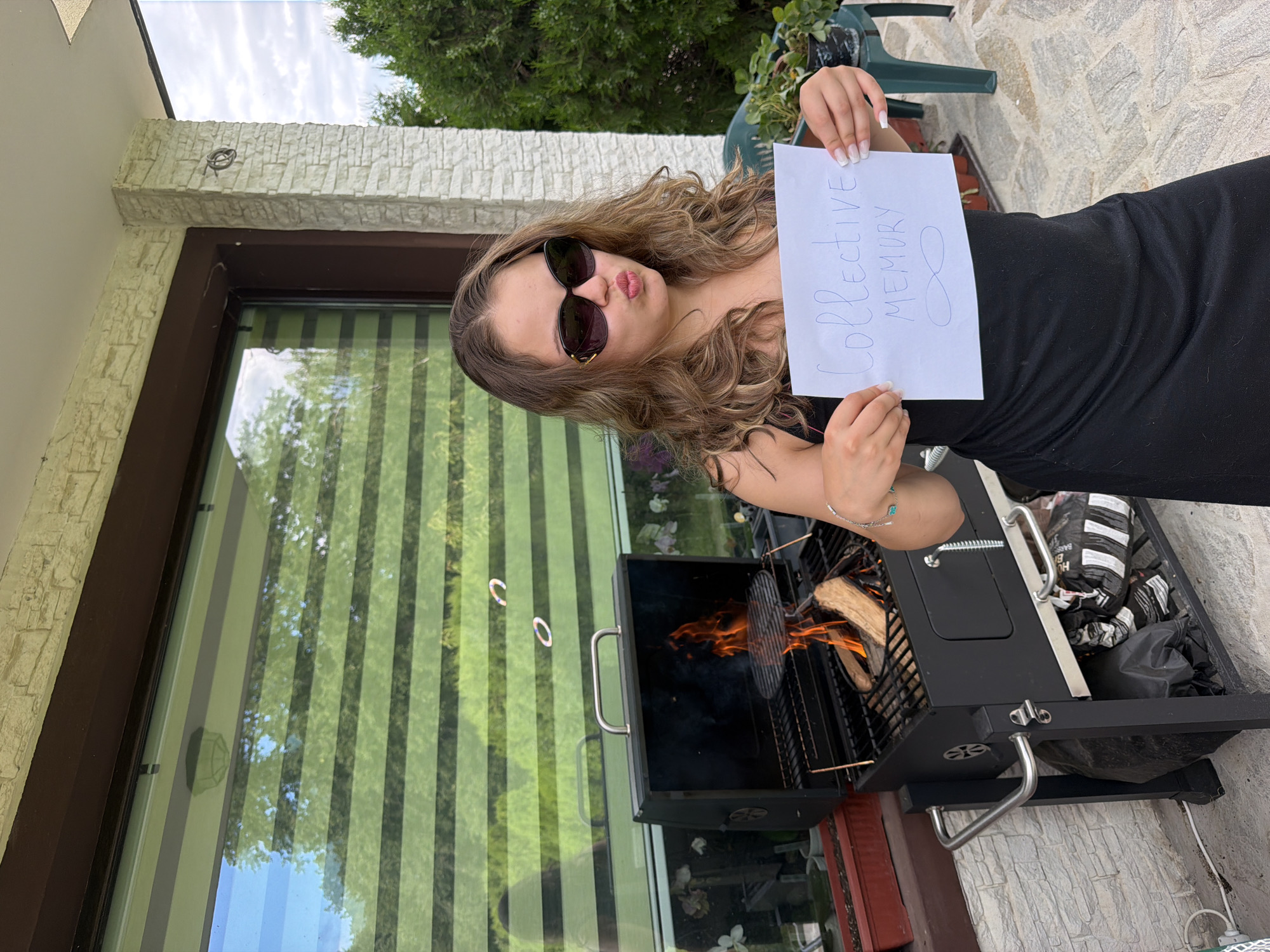 In Priseltsi, Bulgaria, a young woman with brown, wavy hair, wearing sunglasses and a black dress, stands in front of a black barbecue grill. The grill contains burning logs and is positioned on a patio with light-colored stone tiles. Behind the woman and grill is a house with a textured white stone facade and a large window covered by green and white blinds. The woman holds a white piece of paper with "Collective MEMORY" and an infinity symbol drawn on it in blue ink. The scene is outdoors during the daytime, with trees and greenery visible in the background and a cloudy sky.
