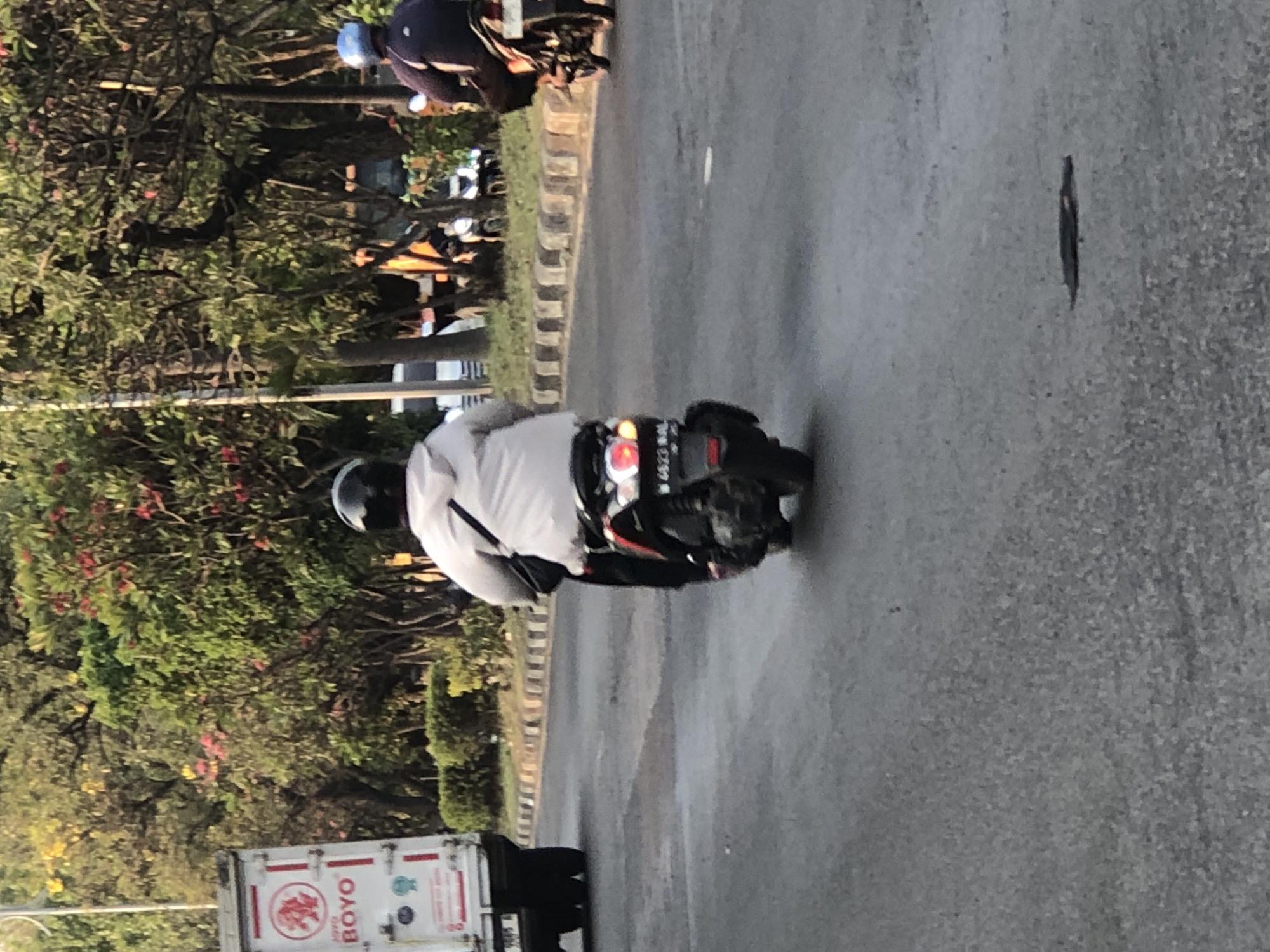 The image shows a street scene in Ngaban, Indonesia, with several vehicles on the road. In the foreground, a person is riding a scooter from behind, wearing a light-colored jacket and a black helmet. The scooter has a license plate visible. To the left, a white truck with "BOYO" written on it is partially visible. In the background, another person is riding a motorcycle, wearing a dark jacket and a blue helmet. Trees and foliage line the side of the road, suggesting an urban or suburban environment. The road surface appears to be asphalt.