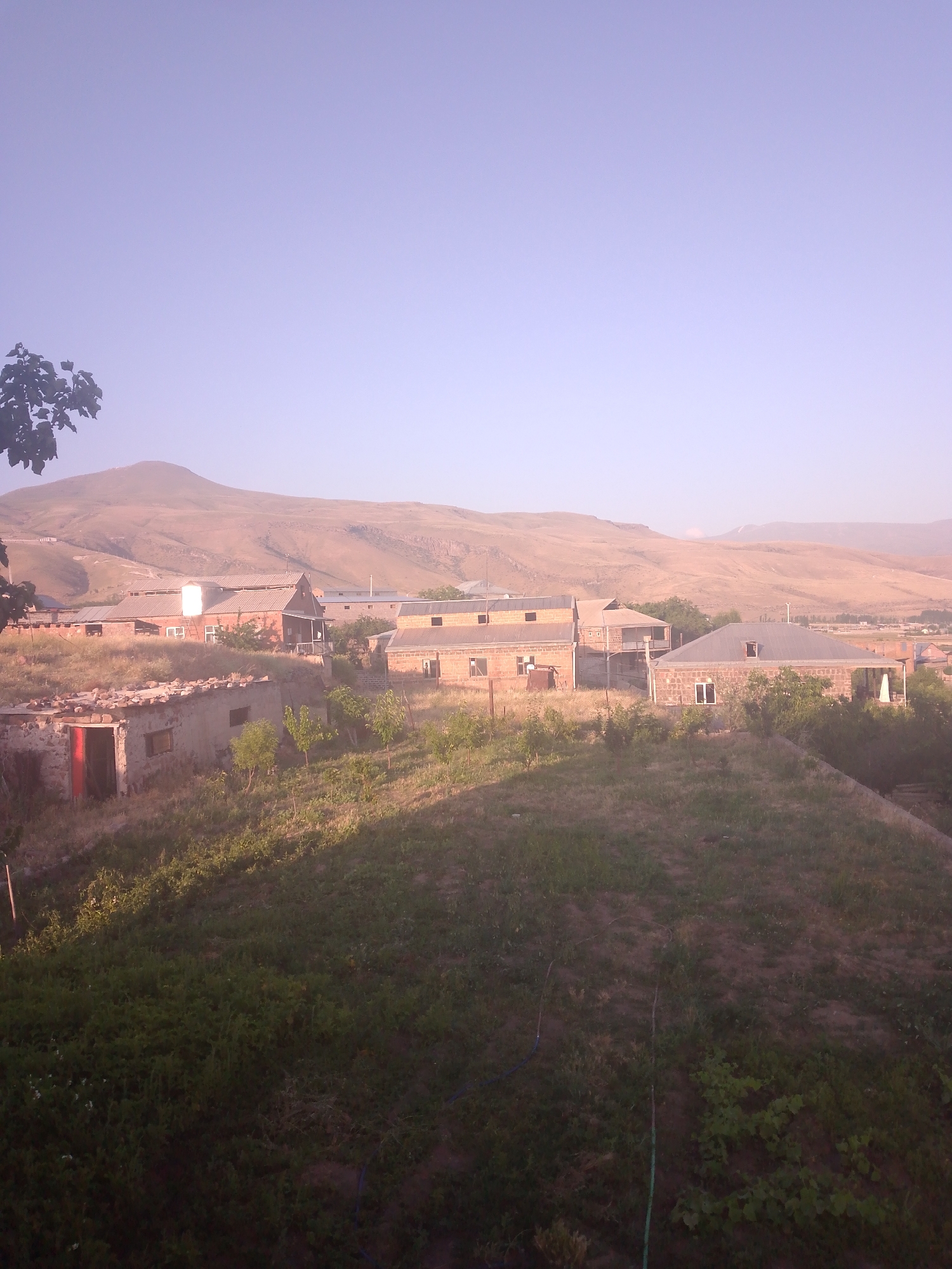 The image depicts a rural scene in Kotayq, Armenia, characterized by a cluster of buildings nestled on a gently sloping hillside, with a large, arid mountain range forming the backdrop. The sky is a pale, clear blue, transitioning to a lighter hue near the horizon. The foreground is dominated by a grassy, uneven terrain with patches of green vegetation and scattered shrubs. A few small, sapling-like trees are visible.
Several structures are present. To the left, a partially ruined, low-lying building with a red-painted door and visible stone and concrete construction stands out. Adjacent to it, a more intact building with a grey metal roof and windows is visible. Further to the right, a row of buildings with grey metal roofs and brick or stone walls are aligned. One of these buildings, slightly larger, has a balcony or porch area. Another building further back appears to be a long, industrial or agricultural structure with a white section on its roof. The architecture suggests a functional, possibly agricultural, settlement rather than a purely residential one.
The overall impression is one of a tranquil, somewhat rustic, and sparsely populated environment, bathed in the soft light of what appears to be early morning or late afternoon. The landscape is characterized by its natural, undeveloped state, with the human-made elements integrated into the existing topography. No people are visible in the image.