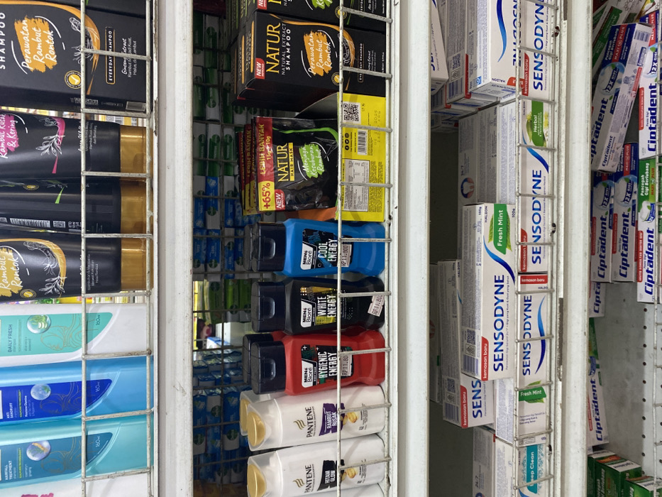 A shelf in a store in Kecamatan Piyungan, Indonesia (latitude: -7.837, longitude: 110.474) with various toiletries and personal care products. On the top shelf are several bottles of shampoo, including "Pantene" and "Rambut Rontok" brands. The middle shelf contains several bottles of hair care products, including "Men's Biore", "Natura", and "Cool Energy" brands. The bottom shelf is stocked with several tubes of toothpaste, including "Sensodyne" and "Ciptadent" brands. The shelves are made of metal with a white background. The lighting is fluorescent. The products are arranged in a neat and orderly manner. The shelves are relatively empty. There are no people in the image.