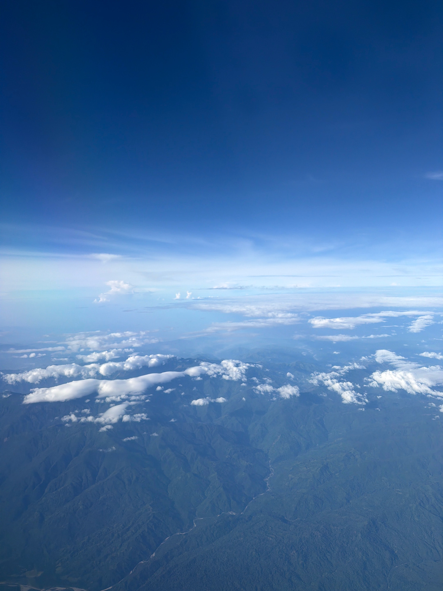 An aerial view captures the vast expanse of a deep blue sky transitioning to lighter hues near the horizon. A layer of fluffy white clouds is scattered across the sky, some appearing as thin wisps and others as more defined formations. Below the clouds, a landscape of lush green mountains is visible, characterized by rolling hills and deep valleys. A winding river or stream is discernible within one of the valleys. The image does not show any identifiable landmarks or structures that would suggest a specific location within Batangas City, Philippines.