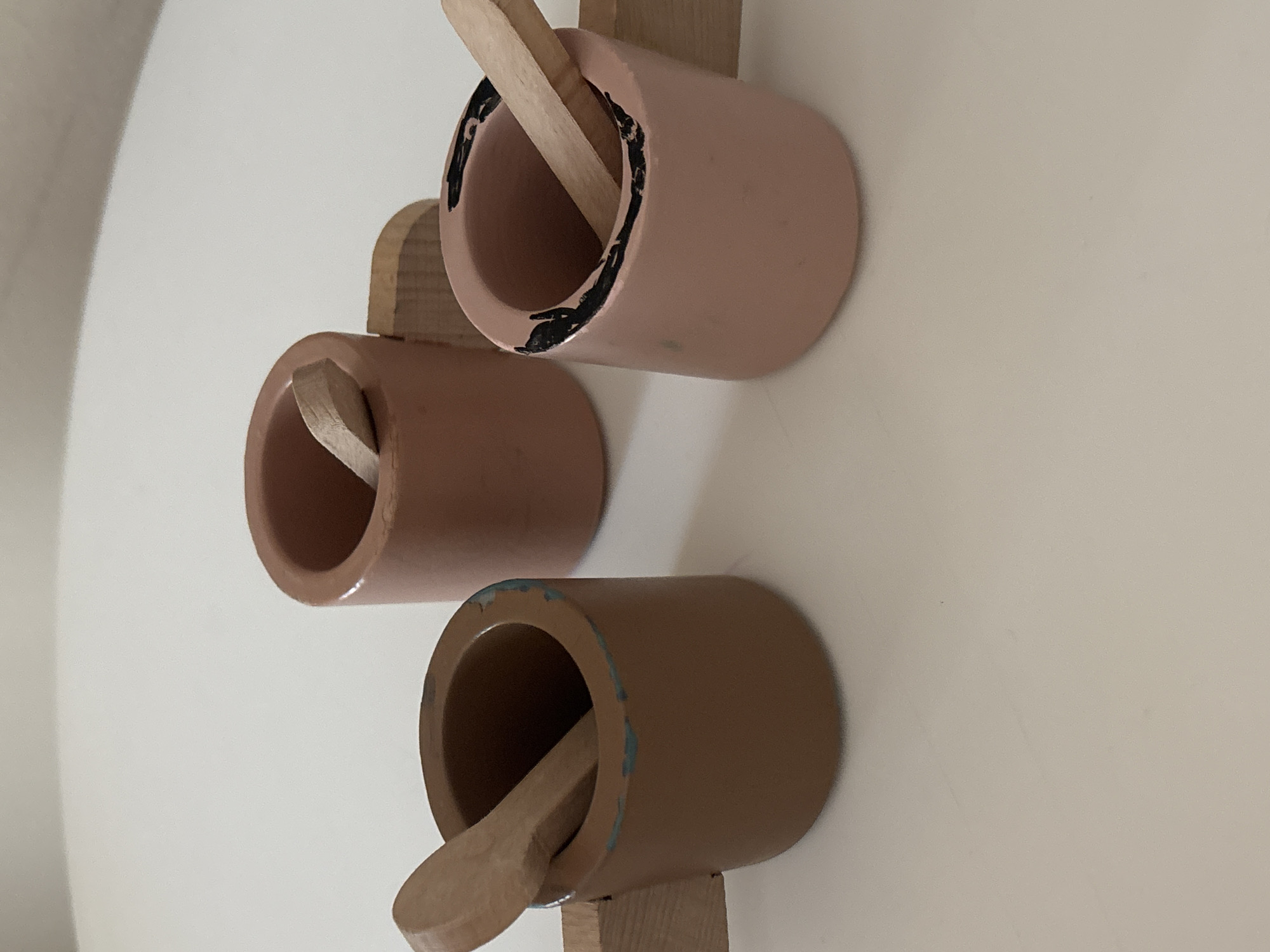 Three wooden toy cups with spoons are arranged on a light-colored, flat surface, possibly a table. The cups are cylindrical with attached handles. One cup is a reddish-brown color and has a wooden spoon inside. Another cup is a darker brown and also contains a wooden spoon. The third cup is a pale pink color with black markings around the rim and has a wooden spoon inside. The lighting suggests an indoor setting with soft illumination. The surface appears to be slightly textured, and there are no other objects or people visible in the frame. The scene is devoid of any contextual clues to place it within Dubai, United Arab Emirates.