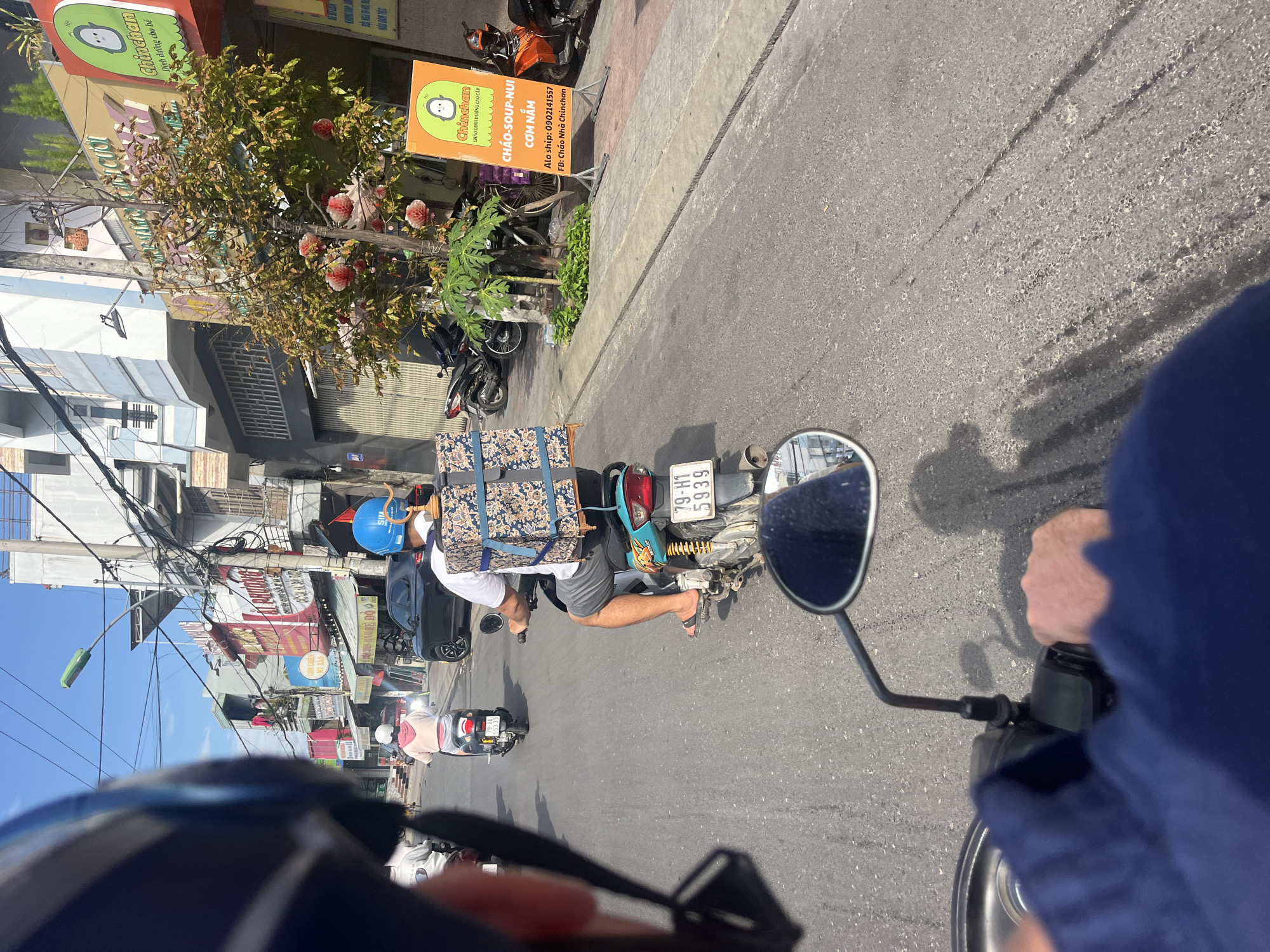 The image captures a vibrant street scene in Bac Nha Trang Ward, Vietnam, viewed from the first-person perspective of someone riding a motorbike. The bright daylight and clear blue sky suggest a sunny day.
In the midground, another person rides a light blue motorbike, moving in the same direction. This rider wears a blue helmet, a white t-shirt, gray shorts, and flip-flops, with their bare legs visible. A sizable package, wrapped in blue fabric with an intricate patterned design, is secured to the back of their motorbike. The motorbike's license plate, "79-H1 5939," is clearly legible.
The right side of the image features a sidewalk lined with buildings, characteristic of Vietnamese shop-houses. Several commercial signs adorn these buildings. Two prominent orange signs for a brand named "Chinchan" are visible. One sign advertises "CHÁO DINH DƯỠNG CAO CẤP" (Premium nutritious porridge) and lists offerings like "CHÁO-SOUP-NUI" (Porridge-Soup-Noodles) and "CƠM NẮM" (Rice Balls), along with contact information: "Alo ship: 0902141557" and "FB: Cháo Nhà Chinchan". The second "Chinchan" sign mentions "Dinh dưỡng cho bé" (Nutrition for babies). A tree with sparse foliage and some decorative artificial pink and red flowers stands in front of these establishments. Overhead, a dense network of electrical wires and cables stretches across the scene.
The road surface is asphalt, showing some tire marks. In the foreground, parts of the photographer's blue sleeve, hand gripping a handlebar, and a rearview mirror are visible, framing the scene. Further down the street, other motorbikes are parked or in motion. The Vietnamese text on the signs and the license plate firmly establish the geographical context.