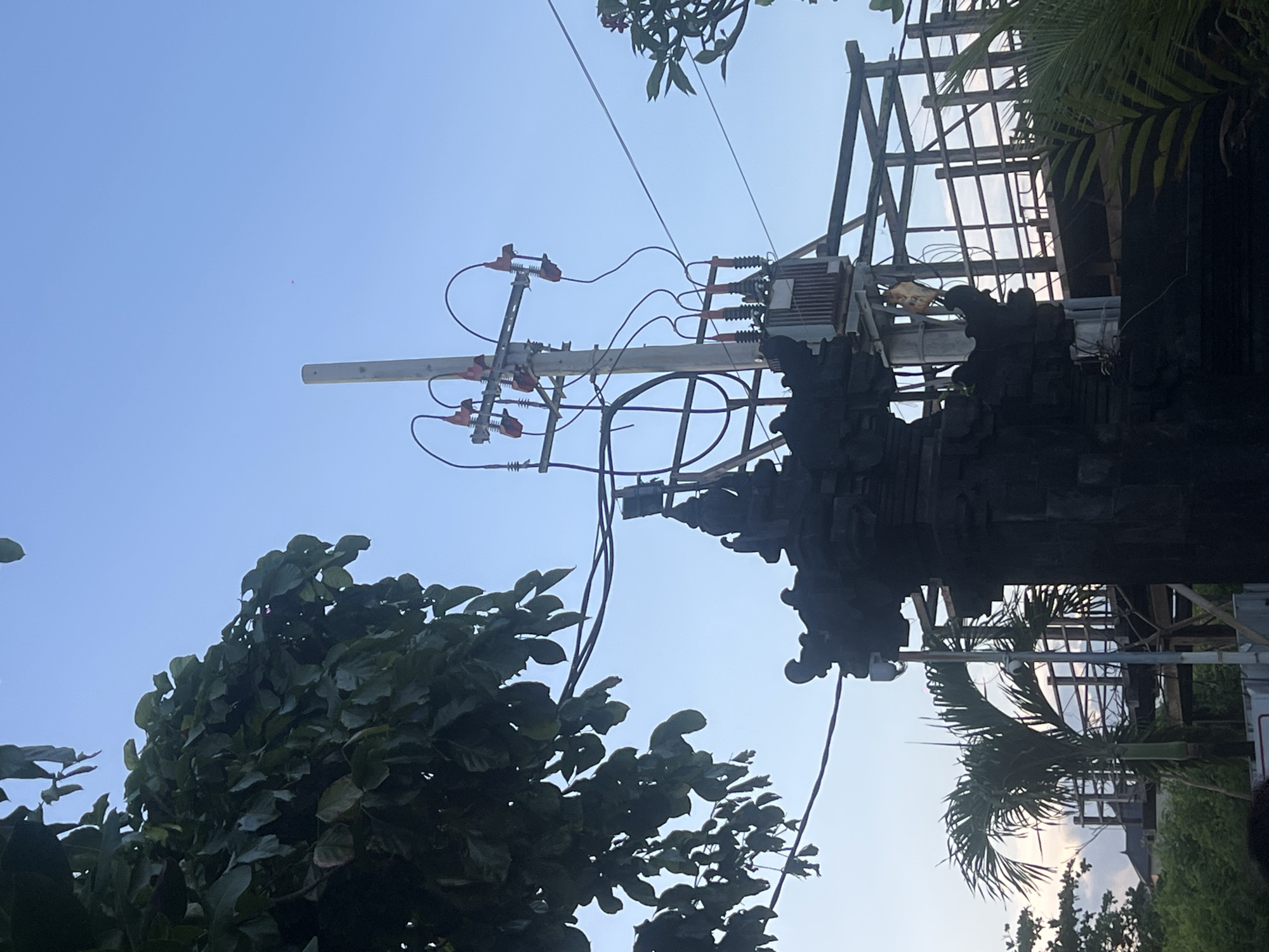 The image shows an electrical pole with a transformer and related hardware, positioned near a Balinese temple structure in North Kuta, Indonesia. The scene is viewed from below, with the blue sky visible in the background. The electrical pole is supported by wooden scaffolding and has several wires extending from it. Lush green trees partially frame the scene.