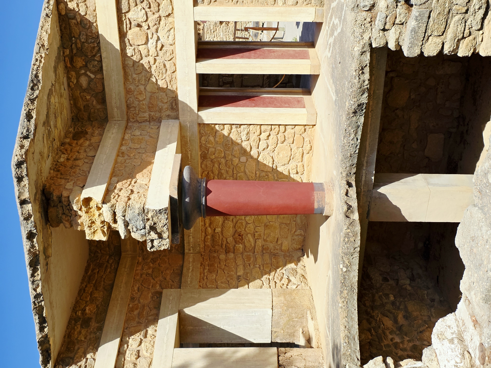 The image shows an interior view of the Palace of Knossos in Heraklion, Greece. The architecture is characterized by rough stone walls, weathered concrete slabs, and a prominent red cylindrical column. The column is topped with a dark, ornate capital and supports a beam and a concrete roof structure. In the background, there are openings framed by columns, some of which are painted with alternating red and cream vertical stripes. Sunlight casts strong shadows across the scene, highlighting the textures of the stone and the geometric shapes of the architecture. The ground is a combination of dirt and flagstones, with some exposed stone foundations visible in the lower part of the image. The sky above is a clear, bright blue. There are no people or animals visible in the frame.