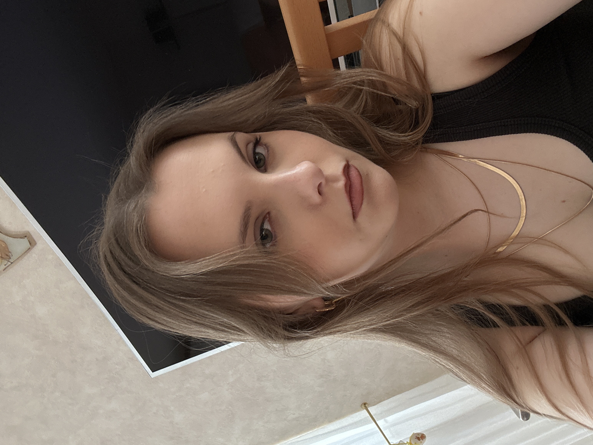 A close-up selfie captures a young woman with fair skin, light brown hair, and a neutral expression. She has green eyes and appears to be wearing subtle makeup, including eyeliner and lipstick. A black tank top is visible, adorned with two gold necklaces. The background includes a black surface, a wooden chair, and a light-colored wall with a textured pattern. There is no indication of the location being in Truchtersheim, France.