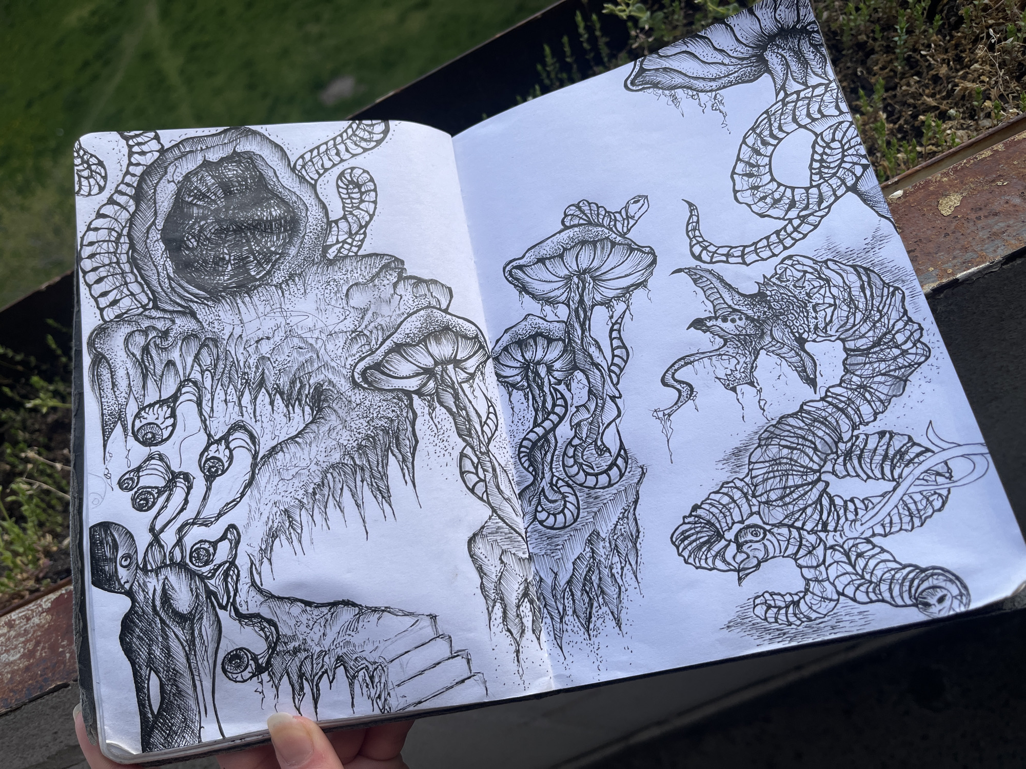 A close-up, eye-level shot features an open sketchbook held by a hand, resting on a weathered, possibly rusty, railing or ledge outdoors in Sofia, Bulgaria. Bright sunlight illuminates the white pages and the intricate pen-and-ink drawings within. The background shows lush green foliage and some dried plants, suggesting a vibrant outdoor setting under clear, sunny skies.
The left page is dominated by a large, menacing hooded figure with a dark, textured void for a face, from which tentacle-like appendages emerge. Below it, a jagged, dripping mass with spiky projections is depicted. Further down, a slender, humanoid figure with multiple eyeballs emerging on stalks from its head and shoulders is drawn. A simple set of stairs is visible at the bottom of the page.
The right page showcases several surreal, mushroom-like structures, some with intertwining tendrils and dripping textures. A long, segmented, serpentine or worm-like creature coils across the top and mid-sections, characterized by intricate patterned scales. Below it, a monstrous, dragon-like head with sharp teeth and spiky features is meticulously drawn, connected to another segmented creature with a bird-like head at its end.
The artwork exhibits a distinct dark fantasy or horror aesthetic, utilizing stippling and detailed line work for rich textures and shading across all subjects. No visible text is present within the drawings or on the sketchbook.