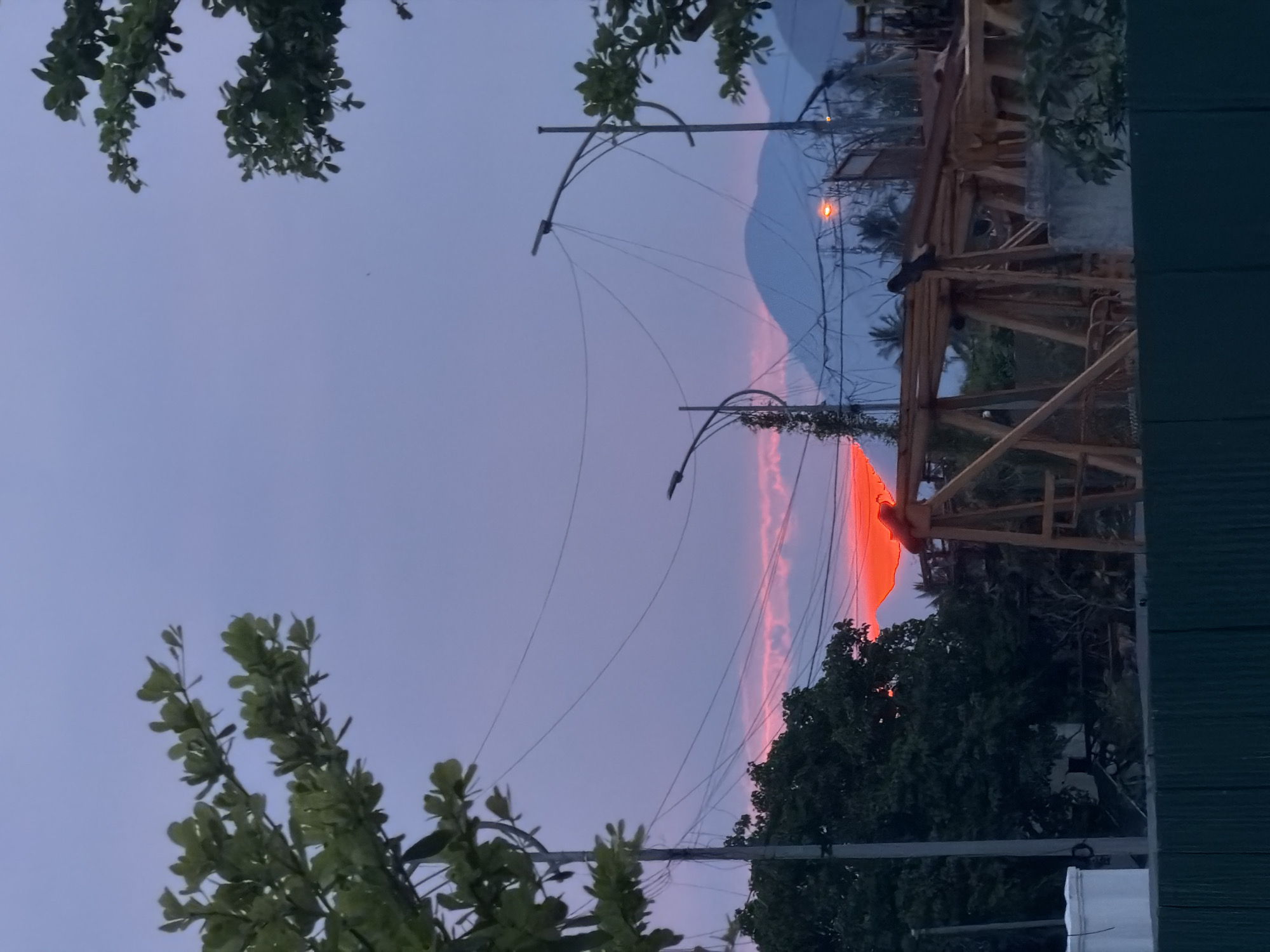 The image depicts a dusky sky over what appears to be a construction site in Nha Trang Ward, Vietnam. A vibrant orange sunset is partially obscured by trees and the silhouette of a large construction crane. Power lines crisscross the sky, strung between lampposts. In the foreground, a dark green corrugated metal fence runs horizontally across the bottom of the frame. The scene suggests the end of a day, with construction activities likely winding down. There are no visible people or animals in the frame.