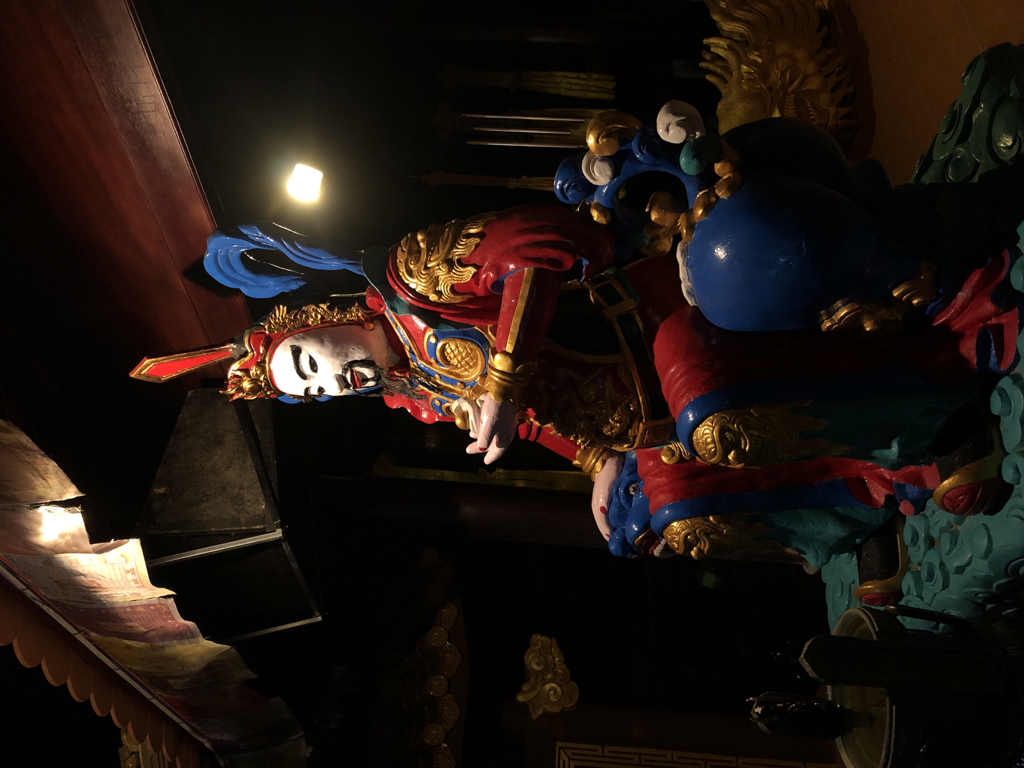 The image shows a colorful, ornate statue of a deity or a historical figure seated on a throne. The figure is wearing a red and gold elaborate costume with a headdress. The statue is in a dimly lit interior, possibly a temple or shrine. Behind the statue, there are wooden beams and decorative elements of traditional architecture. In the foreground, there are some decorative elements in blue and gold, suggesting it's part of a larger altar or display. The overall impression is one of reverence and cultural significance. This statue is located in the city of Mũi Né, Vietnam, likely within a religious or tourist site.