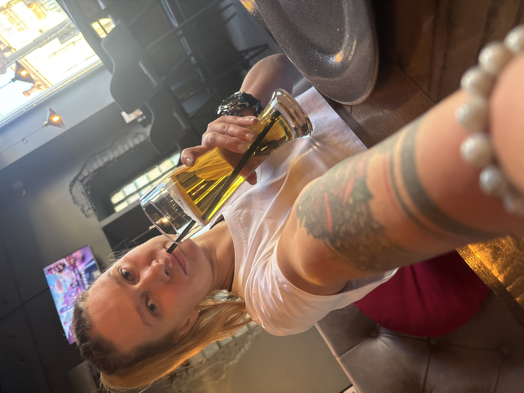 A blonde woman is taking a selfie while seated in what appears to be a restaurant or bar. She is holding a tall glass of beer with a black straw. She has tattoos on her arm. A watch is visible on her wrist. The table has a plate on it, and the woman is seated on a gray button-tufted chair with a red cushion. In the background, there is a TV screen playing an animated show, and a window can be seen. Based on the presence of a TV with animated shows, the location could be a family-friendly establishment. While it's difficult to say with certainty the location is in Perm, Russia, without additional evidence or identifying features specific to that city, the description focuses on what is directly visible and apparent in the photo.