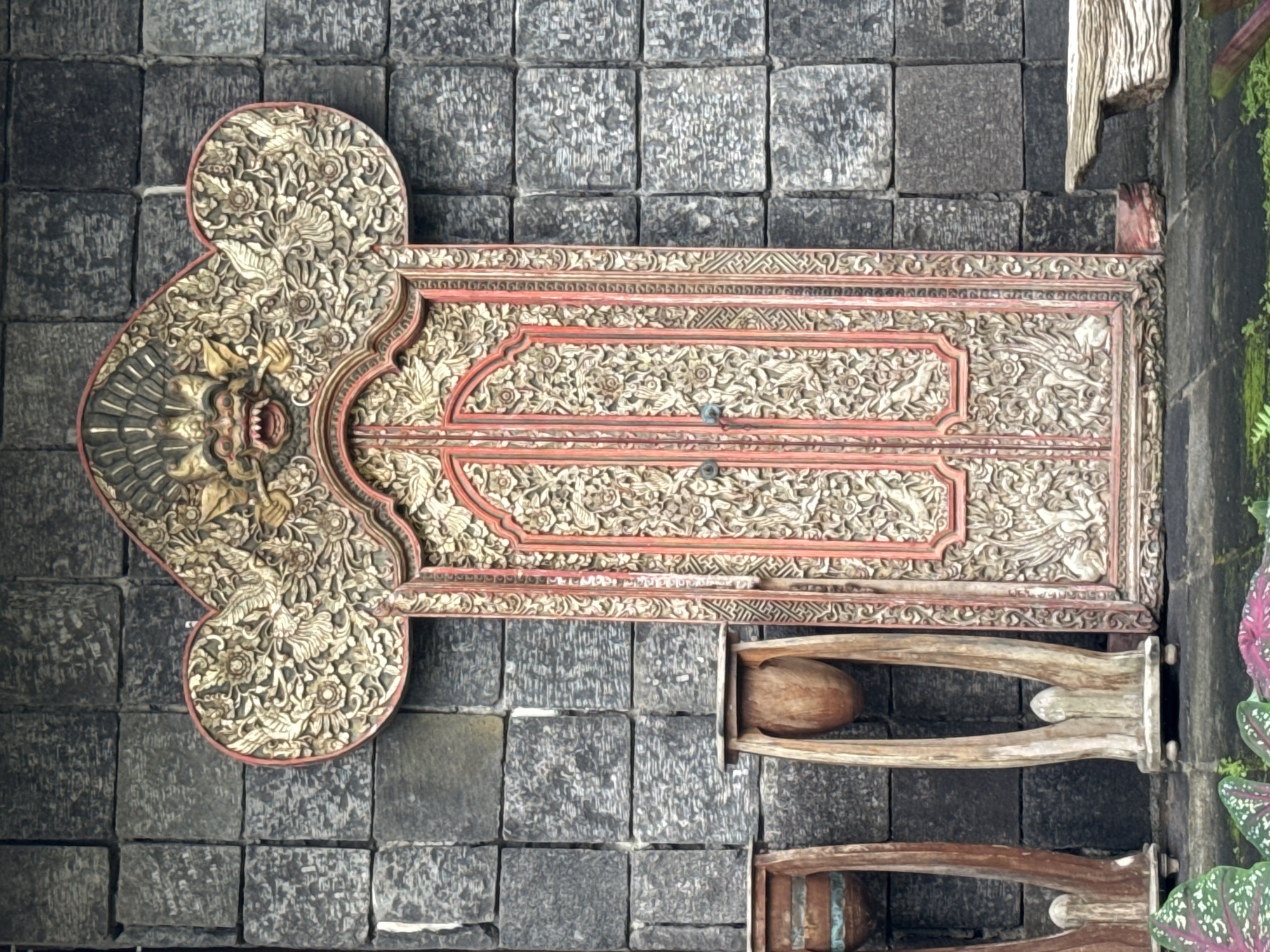 The image displays a highly decorative, intricately carved wooden door or gate, set against a dark grey stone block wall. The door, likely crafted from tropical wood, exhibits elaborate three-dimensional carvings featuring dense floral patterns, scrolling foliage, and depictions of mythical creatures, possibly a stylized Garuda or similar bird-like deity at the top. The carvings are highlighted with shades of gold or light wood, contrasting with deep red accents that outline panels and emphasize details. The door has a distinctive shape, with a broad, rounded, almost heart-shaped upper section, tapering downwards.
Below the main door panels, two wooden stands or furniture pieces are visible. Each features curved wooden legs supporting what appears to be a large, rounded wooden bowl or drum. These items suggest a traditional or cultural setting. To the right, green foliage and plants, including a variety with large, colorful leaves, grow alongside the stone wall, adding a touch of natural vibrancy to the scene.
The style of the carving and architecture strongly suggests a Balinese origin, consistent with the Kuta, Indonesia location context. The dark stone blocks are characteristic of local volcanic stone often used in traditional Balinese construction. The scene is static, devoid of human activity, focusing on the craftsmanship and aesthetic of the architectural elements. There is no visible text.