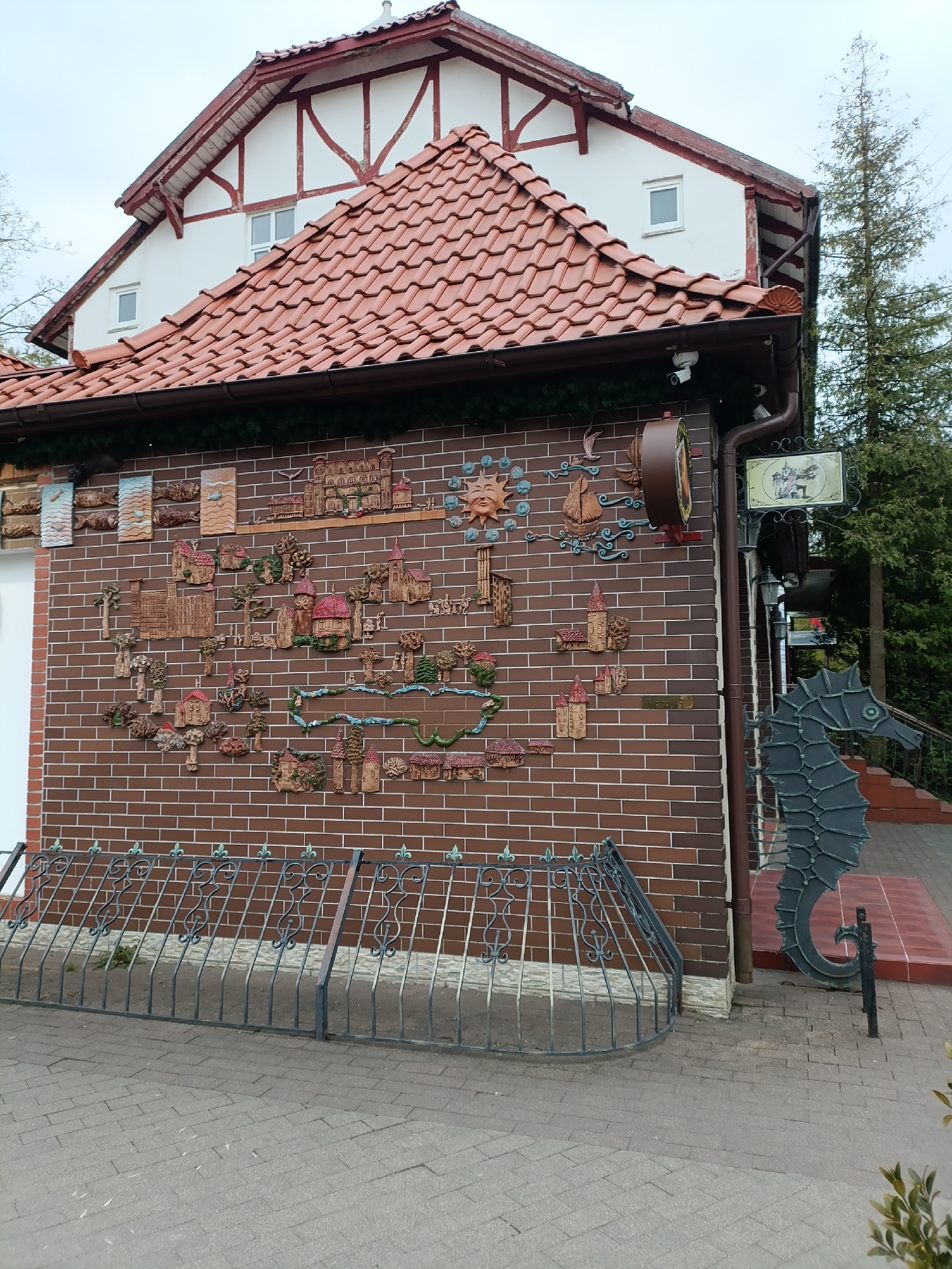 The image depicts a building in Svetlogorsk, Russia. The building has a red-tiled roof and a facade decorated with ceramic reliefs depicting various scenes, including buildings, trees, and aquatic elements. A metal railing with fleur-de-lis accents runs along the base of the wall. To the right, a large, decorative metal sculpture of a seahorse stands next to the building's entrance. A sign hangs above the entrance. The foreground shows a paved walkway.