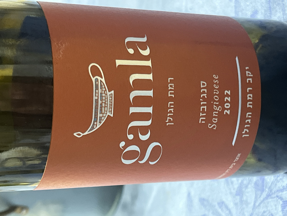 A wine bottle label, predominantly brown, features the name "Gamla" in a large, elegant font. Below the name, in Hebrew, reads "Ramat Hagolan." To the right, "Sangiovese" and the year "2022" are printed. At the top right corner, also in Hebrew, it says "Yekev Ramat Hagolan". A stylized drawing of an ancient oil lamp adorns the label to the left of the name. The bottle itself appears to be green glass.