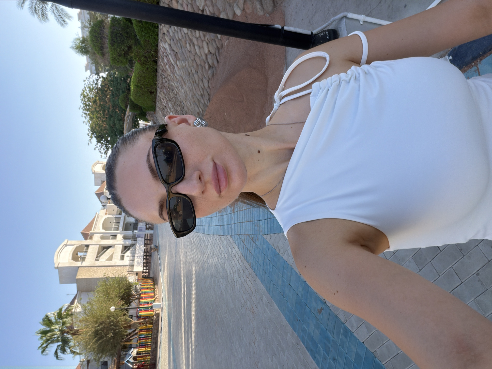 The image shows a close-up, angled selfie of a woman wearing sunglasses and a white top. She has a silver necklace and knot-shaped earrings. The background features a stone wall, palm trees, a multi-story building with arched windows, and a colorful playground structure. The ground is paved with light-colored tiles and a blue pattern. The lighting suggests it is daytime, likely sunny, given the shadows and bright sky. The woman appears to be outdoors, possibly at a resort or hotel in Sharm El Sheikh, Egypt, given the architectural style and vegetation. There is no visible text in the image.