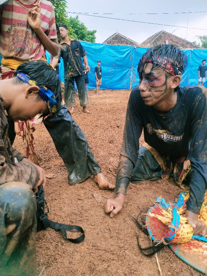 The scene takes place in an open area covered in dirt. There is a blue tarp behind the individuals in the foreground. The area appears to be a performance area, with a young male sitting on the ground with his right leg extended and left leg bent. He is wearing black pants and a black shirt with a white logo and is staring off to the side. His face is painted with black and red markings. His hands are touching the ground, and a yellow, blue, and red drum is lying to his right. He is likely performing a traditional ritual dance. There is a second young male sitting in the foreground with his back to the camera. His legs are extended and his arms are resting on his knees. He is wearing a black and white shirt and black pants. There are two other figures in the background: a young male who appears to be watching the performance and a second young male who is standing to the right of the scene. The location is in Kecamatan Kedawung, Indonesia, with the coordinates -7.494, 111.07. The image shows a group of individuals during a traditional performance.