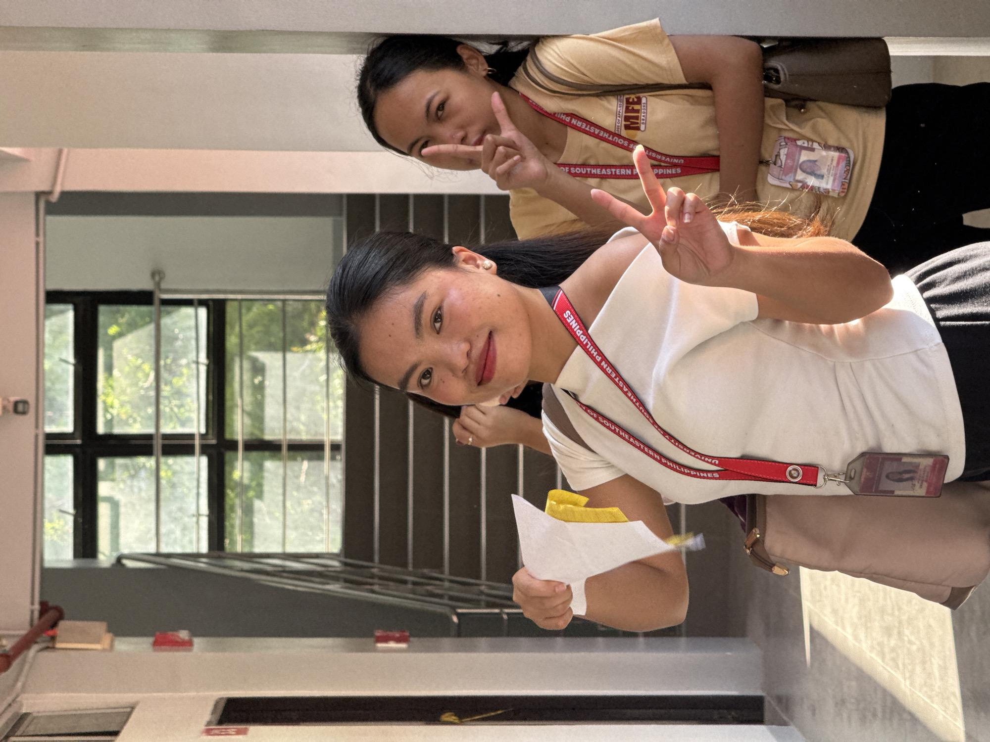 Two young women are posing indoors. One is wearing a white top and holding a paper with yellow items, while the other is wearing a yellow shirt. Both are making peace signs with their hands. They both have lanyards with IDs hanging from their necks. The lanyards read "UNIVERSITY OF SOUTHEASTERN PHILIPPINES." Based on this detail, the location is likely in or near Davao City, Philippines. The building appears to be a university or school building with a window and stairs in the background.