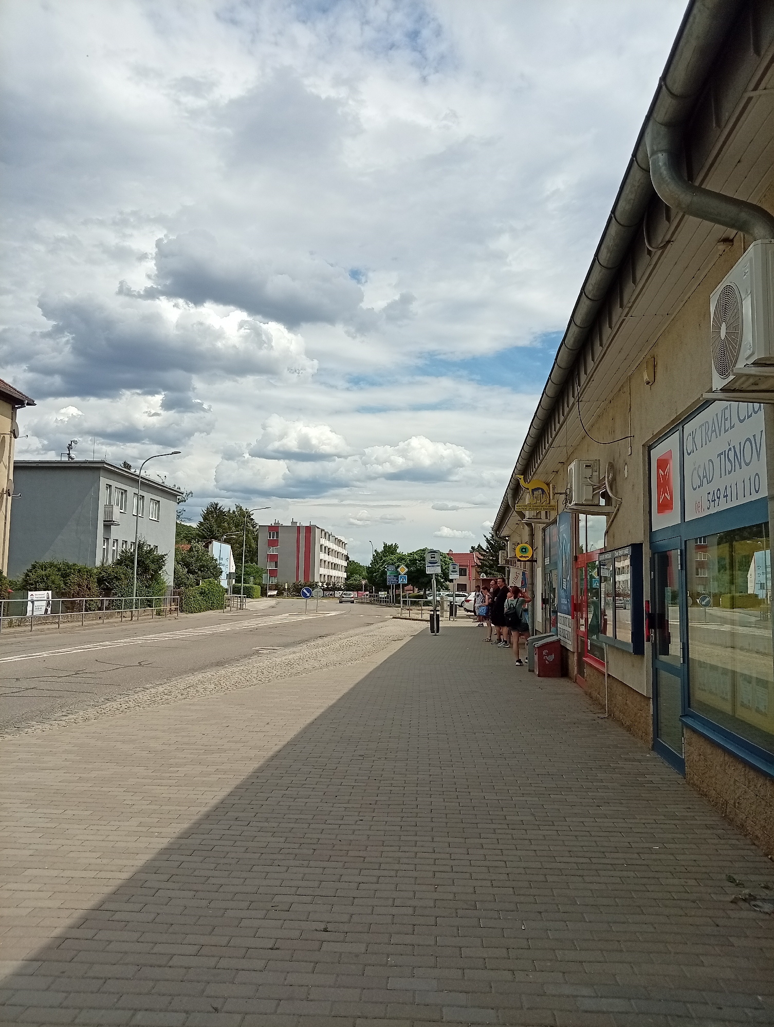 The image shows a street scene in Tišnov, Czechia. The foreground features a paved sidewalk with a strong shadow cast across it, suggesting the sun is high in the sky. On the right side of the sidewalk is a building with a light-colored facade, featuring several windows and doors. Signs are visible on the building, including one for "CK TRAVEL CLUB" and a phone number. An air conditioning unit is mounted on the exterior wall of the building. Several people are standing on the sidewalk in front of the building, appearing to be waiting or browsing. A red bin is also visible on the sidewalk. Across the street, there is a road with pedestrian crossing markings. On the left side of the road, there are multi-story buildings with some greenery and trees. Further down the street, more buildings and trees are visible, along with streetlights. The sky is partially covered with dramatic, dark clouds, indicating potentially changeable weather.
