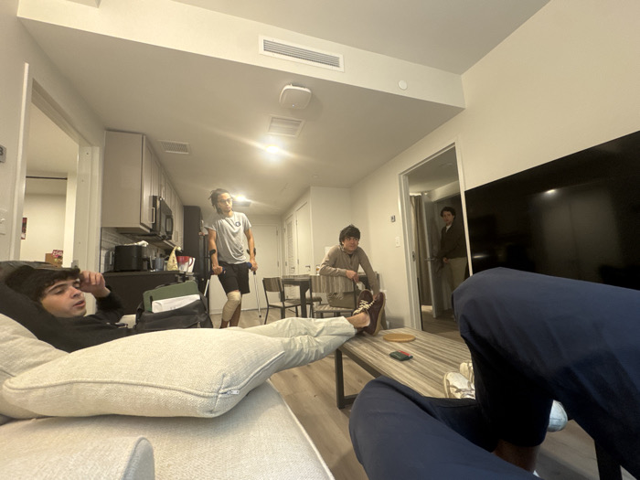The image depicts an indoor scene within a modern apartment in Washington, United States, specifically a combined living room and kitchen area. The perspective is low, from the viewpoint of a person seated on a sofa.
Five individuals are present. In the immediate foreground, the lower torso and legs of a person are visible on the right, clad in dark blue athletic pants and light-colored shoes, with their feet resting on a coffee table. To the left, a young male with dark curly hair and a mustache is reclining on a light-colored sofa, wearing a dark long-sleeved shirt, with his left hand near his head. His legs, covered by light-colored pants, are propped on a cushion.
In the mid-ground, a young male with dreadlocks or long hair is standing in the kitchen area, facing left but looking slightly towards the viewer. He wears a grey t-shirt and dark shorts, utilizing crutches under both arms, and has a brace or bandage on his right knee. To his right, another young male with dark hair is seated at a light-colored dining table, wearing a light brown long-sleeved shirt and dark pants. In the background, partially visible through a doorway on the right, a fifth young male with dark hair stands, wearing a light brown jacket or shirt and light pants, observing the scene.
The environment features light-colored walls, a light-colored wooden or laminate floor, and overhead artificial lighting from ceiling fixtures and recessed lights. The kitchen area includes light-colored upper cabinets, dark lower cabinets, a dark countertop, and stainless steel appliances including a microwave. A large, dark flat-screen television is mounted on the wall to the right. A rectangular wooden coffee table with a striped pattern holds small items including a circular coaster, a green object, and a red object. No direct interactions or specific activities beyond presence in the shared space are discernible.