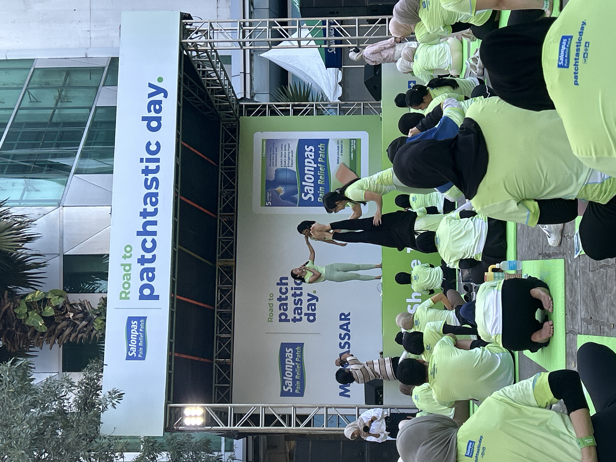 In Makassar, Indonesia, an outdoor event is underway on a stage set up on a paved surface, possibly a street or public square. A large banner above the stage reads "Road to patchtastic day." The stage features a backdrop with branding for "Salonpas Pain Relief Patch." Two women are performing an acrobatic pose on the stage, with one woman in a green athletic outfit assisting the other, who is in a handstand. A third person, also in a green athletic outfit, stands nearby, possibly speaking into a microphone. Below the stage, a group of people, many wearing matching bright green t-shirts and some wearing hijabs, are seated on green mats, participating in an activity. A water bottle and a tablet are visible among the seated participants. To the right of the stage, a white tent is visible, and beyond that, a modern building with a glass facade rises in the background. The lighting suggests it is daytime.