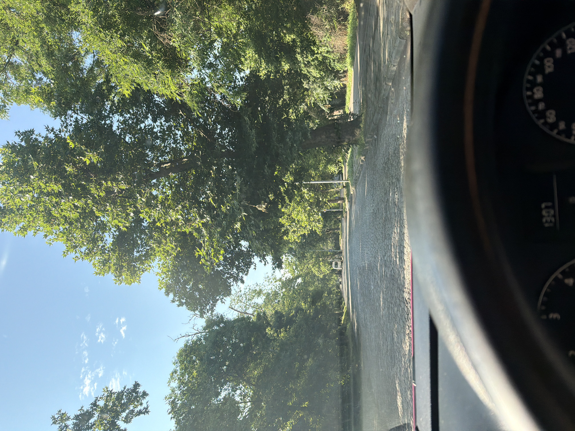 The image captures a perspective from inside a vehicle looking forward through the windshield on a bright, sunny afternoon. The lower portion of the image shows the car's dark dashboard and part of the steering wheel, with the digital clock displaying "13:07".
The central and upper parts of the image reveal a long, straight avenue paved with cobblestones, stretching into the distance. Both sides of the street are densely lined with mature, deciduous trees, likely plane trees, with vibrant green foliage that forms a thick canopy overhead, casting dappled sunlight and shadows on the road. Sidewalks are visible along the tree lines, with some streetlights interspersed among the trees. In the far distance, a white van is visible on the right side of the road. The sky above the tree canopy is clear blue with a few scattered white clouds. The overall scene conveys a tranquil, verdant urban environment.
This specific location is **Aleko Konstantinov Street in Plovdiv, Bulgaria**. This street is famously characterized by its distinct cobblestone pavement and the majestic, dense alley of mature plane trees that create a recognizable green archway, providing shade and a unique aesthetic.