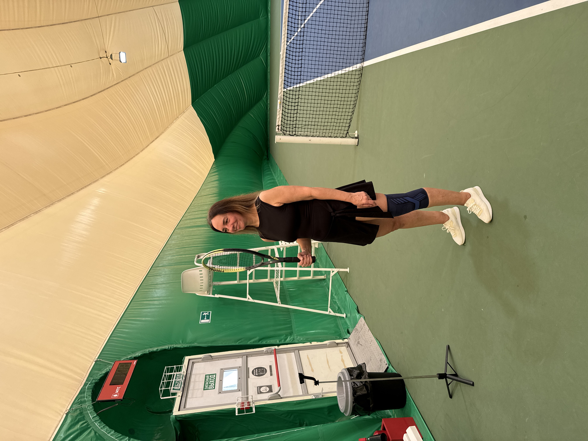 A woman stands on an indoor tennis court in Nizhny Novgorod, Russia, holding a tennis racket. She is wearing a black dress, white sneakers, and a blue compression sleeve on her leg. The tennis court features a blue and green surface, a net, a referee's chair, an exit door, and a scoreboard. The building has a green and beige domed ceiling.