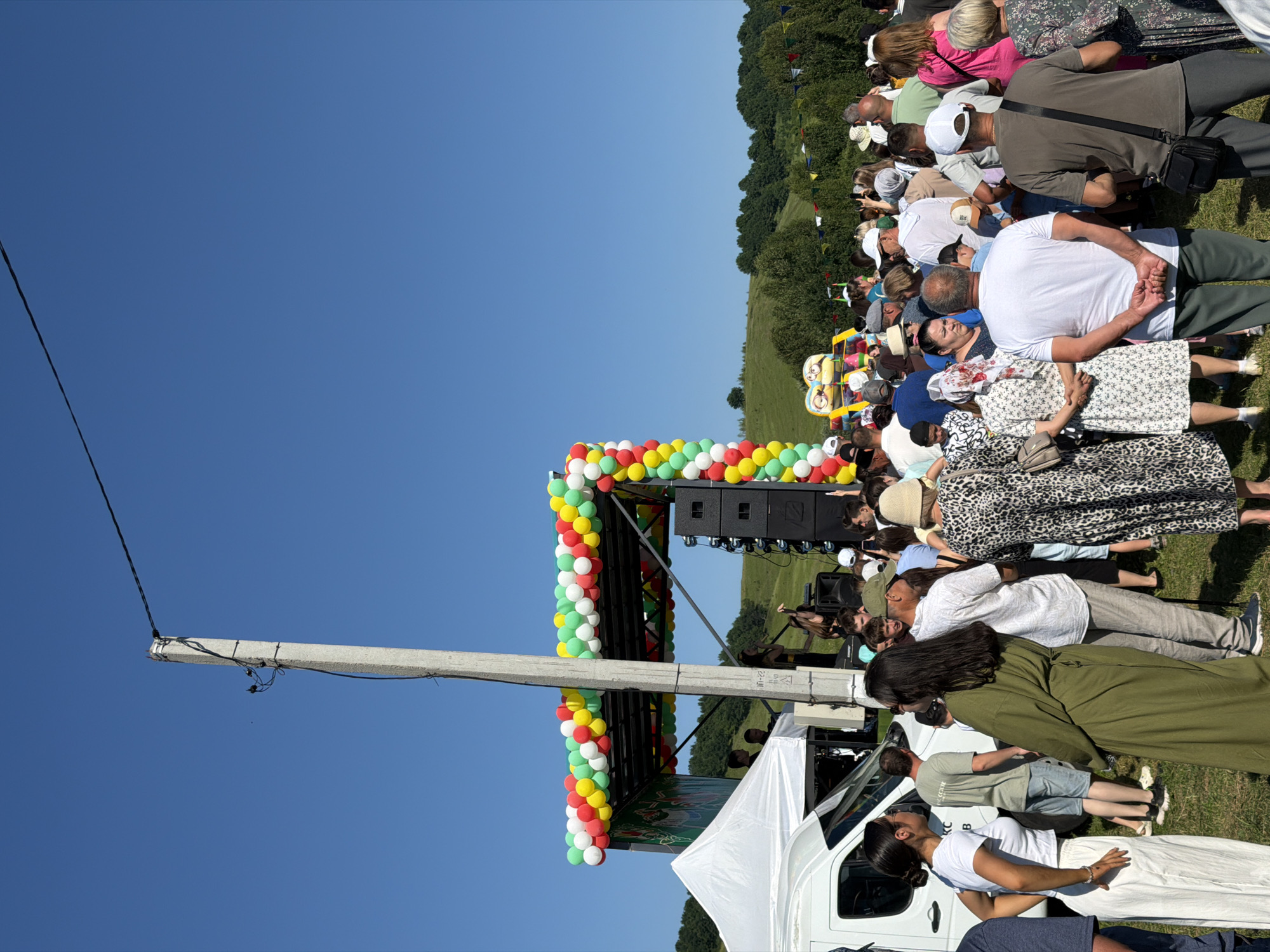 An outdoor daytime scene depicts a large gathering of people attending an event, possibly a festival or concert, on a grassy hillside under a clear blue sky. A stage, decorated with colorful balloons in red, yellow, green, and white, is set up with sound equipment. Several individuals are visible on the stage, including a female performer. A white tent and a white van are situated near the stage. The crowd consists of people of various ages, many of whom are looking towards the stage. The environment is characterized by rolling green hills and scattered trees in the background. A tall concrete utility pole with wires is prominently positioned in the foreground, slightly off-center to the left. The overall atmosphere appears festive and public.