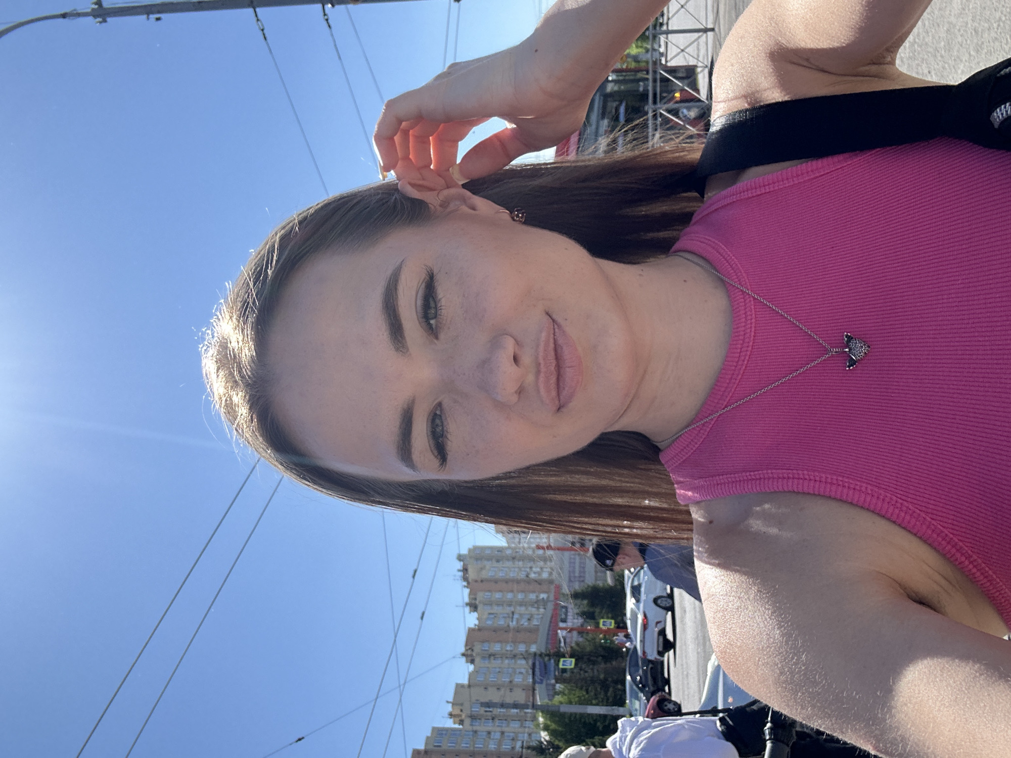 In an outdoor, urban setting under a bright, sunny sky, a young woman is captured in a close-up selfie. She has long, brown hair and is wearing a pink ribbed tank top and a silver necklace with a dark pendant. Her right hand is raised, touching her ear. The background shows a clear blue sky with overhead wires, buildings, trees, and several cars on a street. Some individuals are visible in the background, including a person in a white jacket on the left and another person in a dark shirt and cap. The scene appears to be a street or intersection within a city.