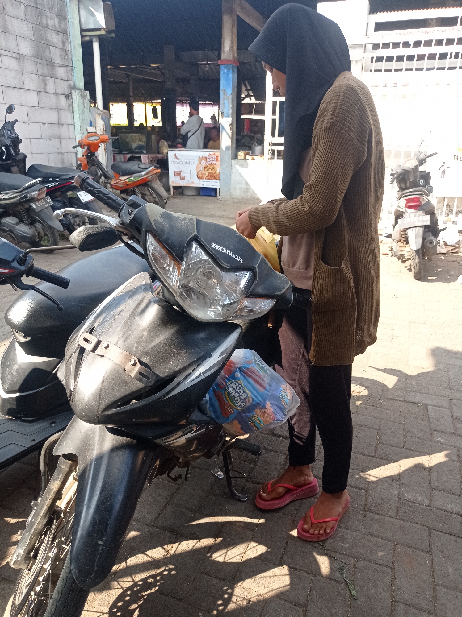 A young woman wearing a black hijab, a brown cardigan, and dark pants stands next to a parked black motorcycle in an outdoor market setting in Tajinan, Indonesia. She is holding a yellow plastic bag in her hands and appears to be interacting with it. The motorcycle is a Honda model, visible with its headlights and front fender. A plastic bag containing colorful packaged snacks is attached to the front of the motorcycle. Other motorcycles are parked in the background, along with stalls and structures characteristic of a local market. Sunlight casts distinct shadows on the tiled ground.