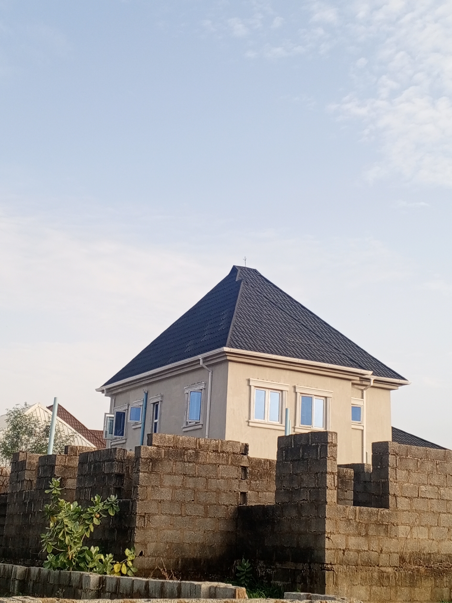 A two-story house with a dark shingled roof sits behind a partially constructed brick wall. The house has light-colored exterior walls and multiple rectangular windows with white frames, some of which are open. A small tree or shrub is visible in front of the wall. The sky is pale blue with some wispy clouds. The location is Unknown, Unknown.