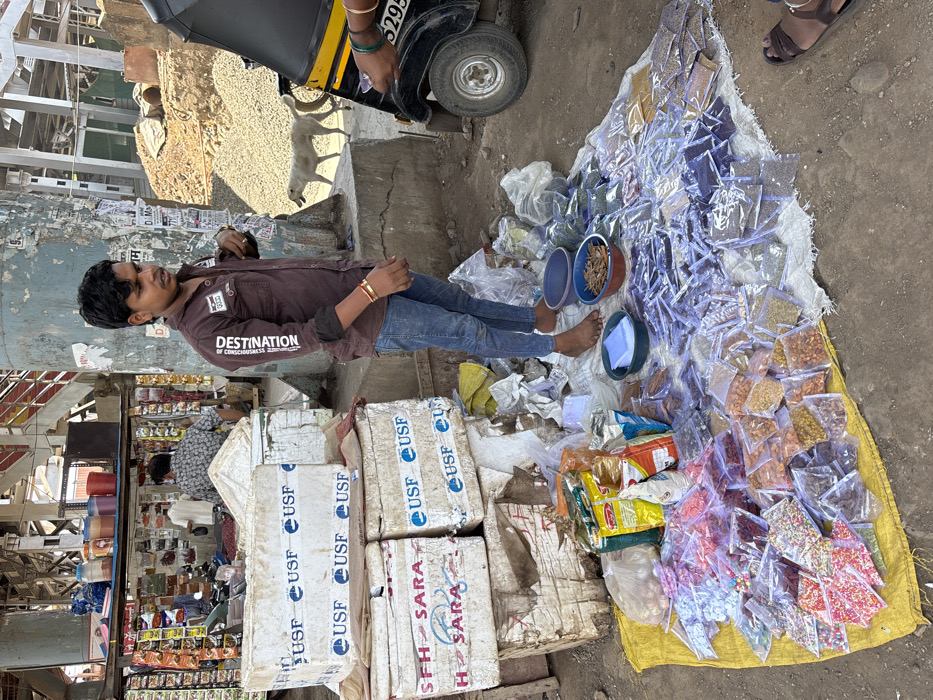 This image depicts an outdoor street vending scene in Ulhasnagar, India, illuminated by bright daylight. A young adult male, barefoot, wearing a brown long-sleeved collared shirt with "DESTINATION" text on the sleeve and blue jeans, stands central, functioning as a vendor. Gold-colored bangles are visible on his right wrist. His street stall is arranged on the dirt ground, featuring a yellow tarp and a white fabric upon which numerous clear plastic pouches containing various dry goods, including spices, grains, nuts, dried fruits, and colorful sprinkles, are displayed. Several larger packaged bags and two plastic bowls, one blue and one reddish-brown containing small items, are positioned near his feet. To the vendor's left, a stack of four white insulated boxes, marked with "USF" and "SARA" branding, is visible. On the far right, the front-right section of a black and yellow auto-rickshaw is partially visible, displaying "3295" on its license plate, with a person's hand, adorned with green bangles, extended outward, possibly in an interaction. In the mid-ground, a tall concrete pillar, covered in distressed paint and various adhesives, serves as a prominent vertical element. Behind the pillar, a secondary vendor stall, displaying packaged items and colorful containers, is attended by two male individuals. A light-colored dog traverses the dirt path in the mid-background. The upper background reveals an elevated metal structure, likely a railway or bridge, with visible support beams, alongside an area of disturbed earth or gravel.