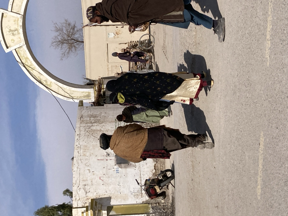 This image depicts an outdoor street scene in Kandahar, Afghanistan, under clear daylight conditions with strong shadows. The foreground shows three individuals on a paved surface. On the left, a person wearing a brown shawl, dark clothing, and a dark turban is seen from the back, walking away. To their right, another person, also seen from the back, wears a dark patterned head covering and outer garment over a cream-colored dress with maroon trim, also walking away. On the right, a bearded man, facing left, wears a brown shawl over a light blue tunic and dark blue pants; he holds a small clear bag containing nuts or seeds.
In the mid-ground, another individual in green clothing walks away. Further back, beneath a weathered white archway, a person rides a bicycle, and another individual in military-style fatigues is partially visible. To the left, a cracked and weathered white wall with markings, including "303," is visible, next to which a motorcycle with luggage is parked on its kickstand. Bare trees are present in the background. A dark cable runs across the upper part of the scene.