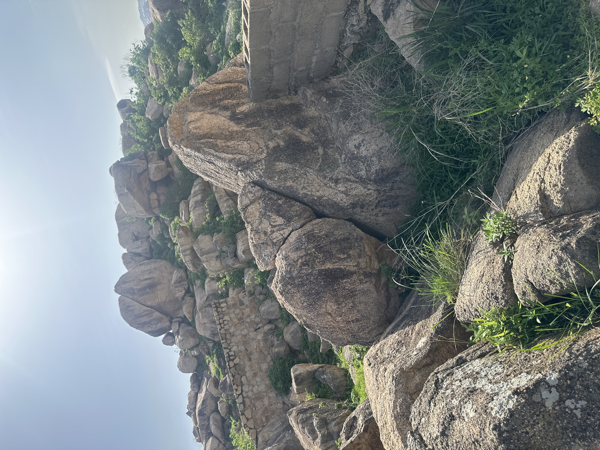 The image depicts a rocky, hilly landscape with scattered vegetation and some man-made structures. The foreground is dominated by large, weathered boulders. In the midground, a low stone wall, appearing aged and partially ruined, is visible among the rocks and greenery. Further back, more rocks and dense green bushes cover the slope. A partial view of a concrete block structure is present on the right side of the image, suggesting human presence or past construction. The sky above is bright and hazy, with the sun clearly visible, indicating a daytime scene. The overall impression is of a natural, somewhat rugged environment on the outskirts of a settlement. This type of landscape, characterized by granite rock formations and interspersed vegetation, is characteristic of areas around Jos, Nigeria, known for its geological features.