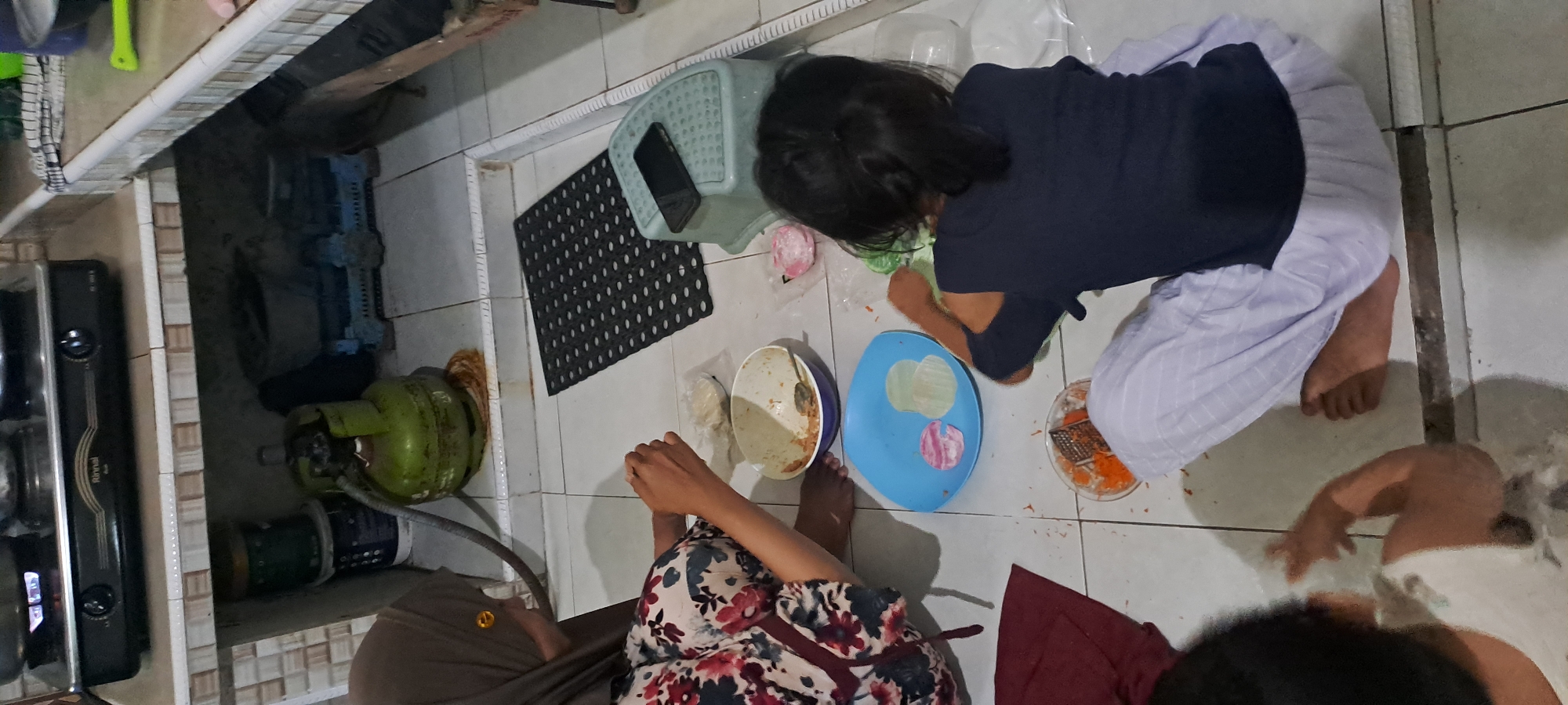 In a home kitchen located in Cemandi, Indonesia, two children and an adult are engaged in food preparation on a tiled floor. The adult, wearing a floral-patterned top and a brown hijab, is on the left, seated on the floor. The two children are also seated on the floor. One child, wearing a dark blue shirt, is in the center-right, actively grating carrots into a bowl. The other child, partially visible on the far left, is wearing a white shirt.
In the foreground, various food items and utensils are scattered. A blue plate contains colorful circular food items, likely wrappers or dough, with some grated carrot on it. A bowl filled with grated carrots and a spoon sits to the left of the blue plate. A red cloth lies to the left of the adult. Behind the children, a black mat is placed on the floor, with a smartphone resting on a small green stool to its right.
The background reveals the kitchen's lower section. A green gas cylinder is prominently placed next to stacked containers. A black gas stove with visible flames is on a shelf above, indicating active cooking. The kitchen appears functional, with basic household items and cooking apparatus visible. The lighting suggests an indoor setting, possibly during the day.