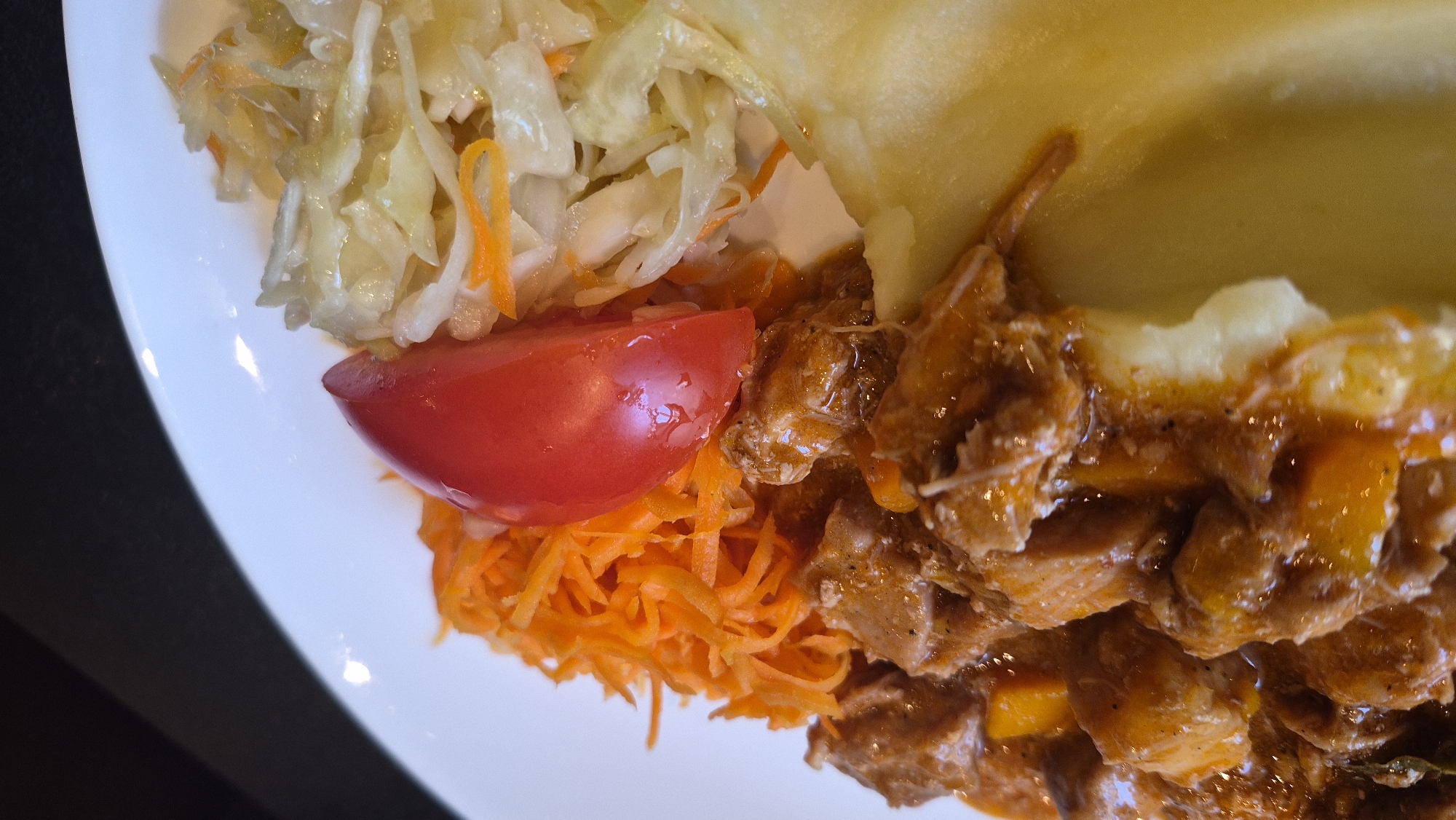A close-up, top-down, vertically oriented shot shows a white plate with food. On the plate, there is a serving of mashed potatoes to the right, topped with a slice of tomato and a side of coleslaw. To the left of the coleslaw and tomato is a pile of shredded carrots. Below the carrots and tomato, a hearty portion of stewed meat with visible pieces of carrots is served over mashed potatoes. The background is dark, with a hint of a black surface on the upper left. The food appears to be from a restaurant in Daugavpils, Latvia.
