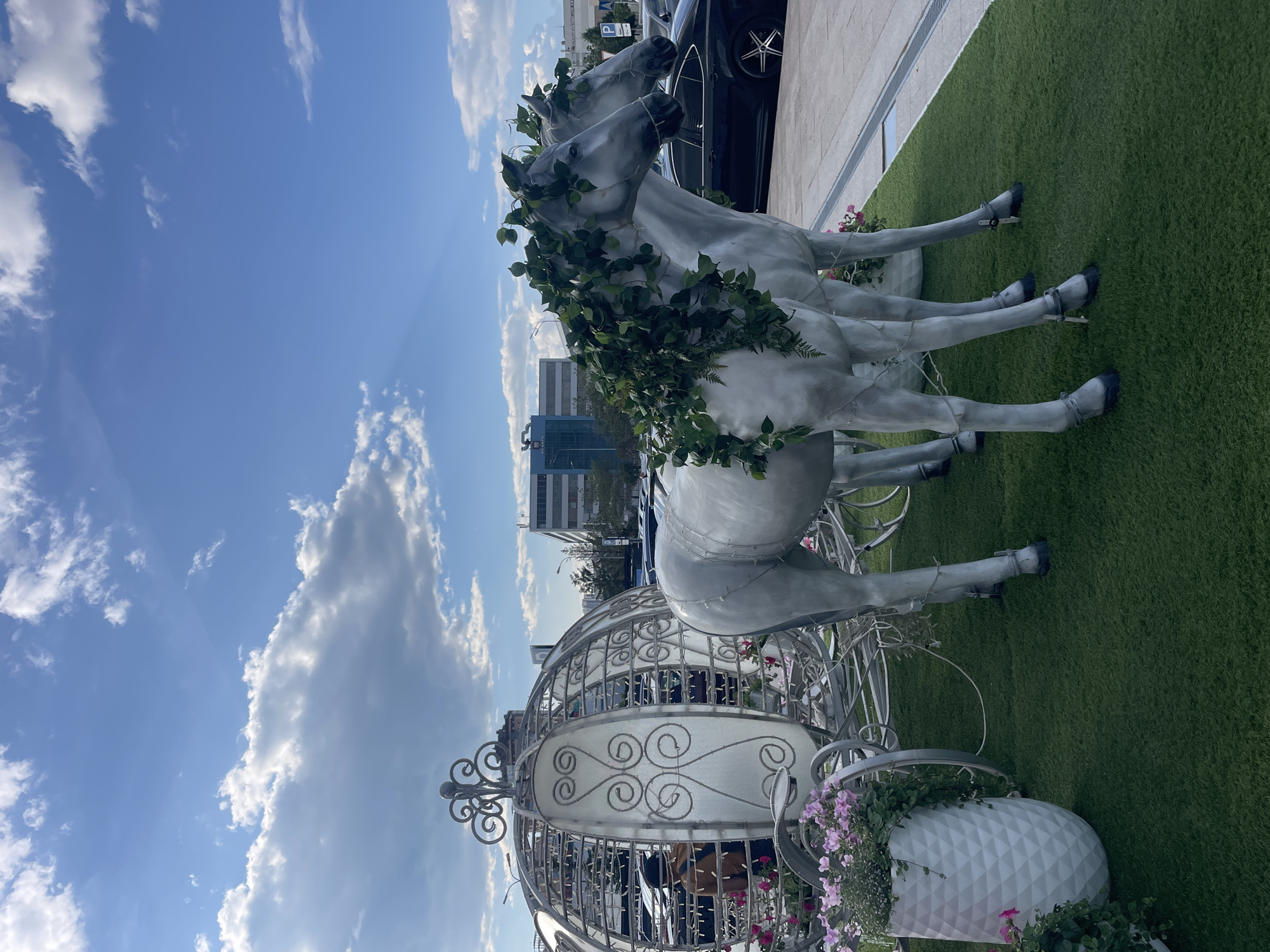 The image depicts a decorative display featuring a white Cinderella-style carriage pulled by two white horse statues. The horses are adorned with green foliage. The display is set up on a patch of artificial grass, with a white planter filled with pink flowers to the left of the carriage. In the background, a parking lot with cars and several modern buildings are visible under a blue sky with scattered clouds. The scene appears to be outdoors, possibly in a public space or event venue. The location is identified as Vysokaya Gora, Russia.