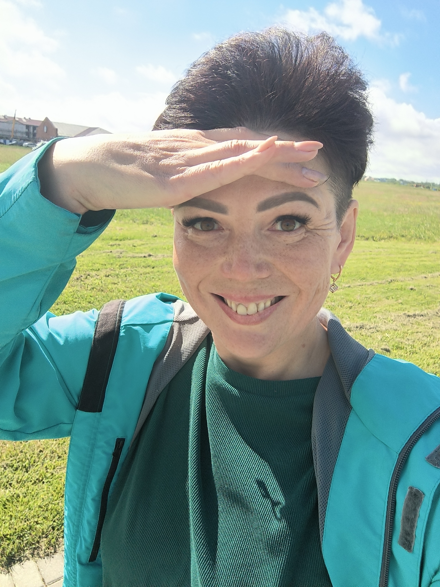 A woman stands in a grassy field under a bright, cloudy sky, possibly in Laskino, Russia. She is taking a selfie and shielding her eyes from the sun with her left hand. The woman has short, dark hair, freckles, and is wearing a teal jacket over a dark green ribbed shirt. She is smiling and has visible makeup, including mascara and lipstick. A small, geometric earring is visible on her right ear. The background shows a green, rolling hill with some buildings in the distance. The lighting suggests it is daytime, and the presence of a jacket implies it is not extremely hot weather.