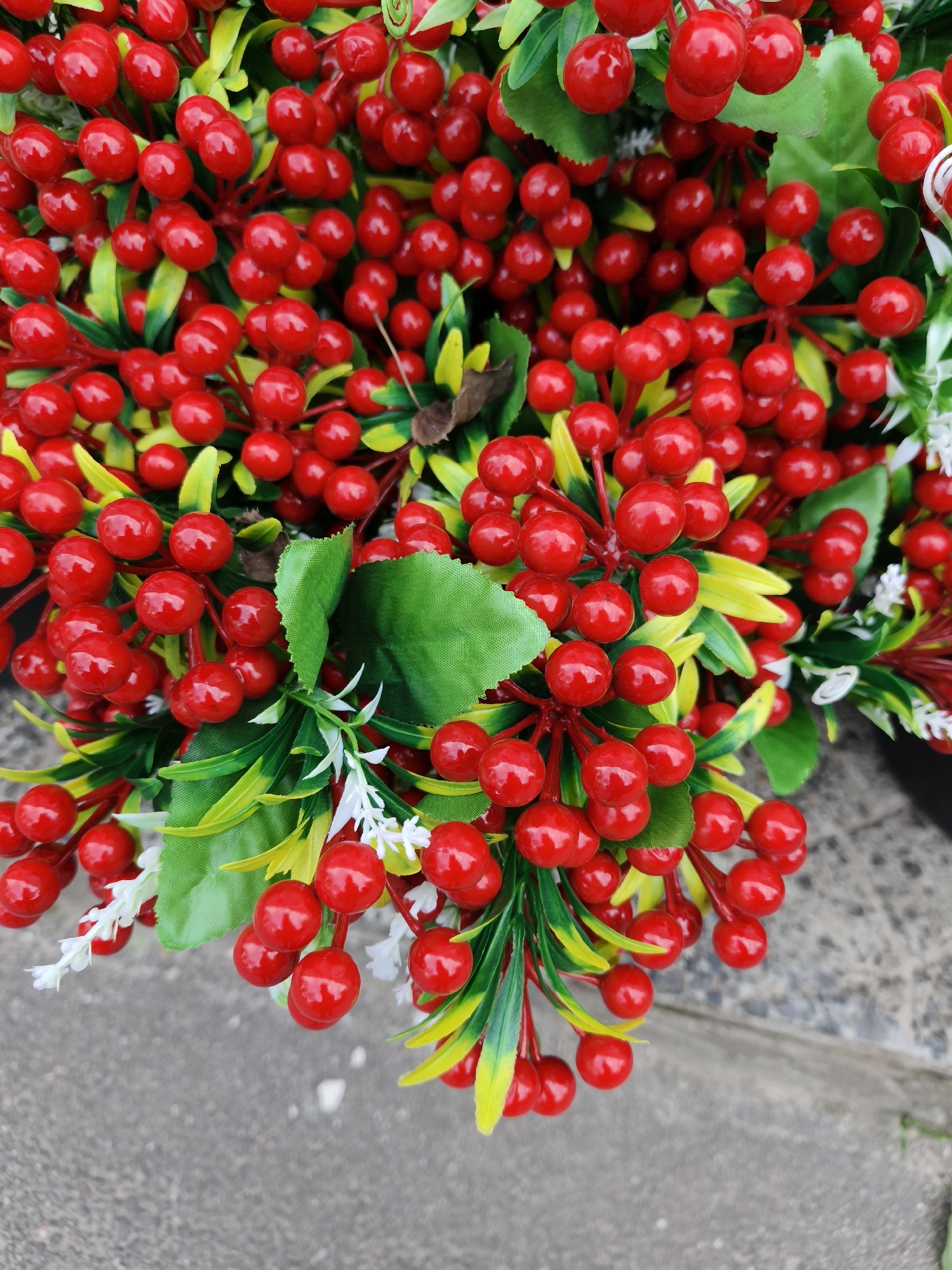 The image presents a close-up, top-down view of a vibrant arrangement of artificial red berries, green leaves, and small white flowers. The central and most prominent feature is a dense cluster of numerous bright red, shiny berries, which appear to be made of plastic or a similar synthetic material. These berries are uniformly round or slightly oval and are attached to thin, reddish-brown stems.
Interspersed among the berries are various artificial leaves. Some are broad and a consistent deep green, while others are more slender and exhibit a gradient from bright green to a yellowish-green, adding textural and color variety to the foliage. Small, delicate, white artificial flowers, resembling tiny starbursts, are scattered throughout the arrangement, providing a subtle contrast to the dominant red and green tones.
The decorative elements fill the majority of the frame, suggesting a full and lush display, possibly part of a bouquet, wreath, or another festive decoration. The background, visible at the bottom and along the edges, is a textured grey surface, likely concrete or asphalt, with minor tonal variations and faint lines. This suggests the arrangement is placed outdoors or on a hard ground surface in Kryvyi Rih, Ukraine. The lighting is bright and even, highlighting the glossy finish of the berries. There is no discernible activity taking place, nor is any text visible in the image.