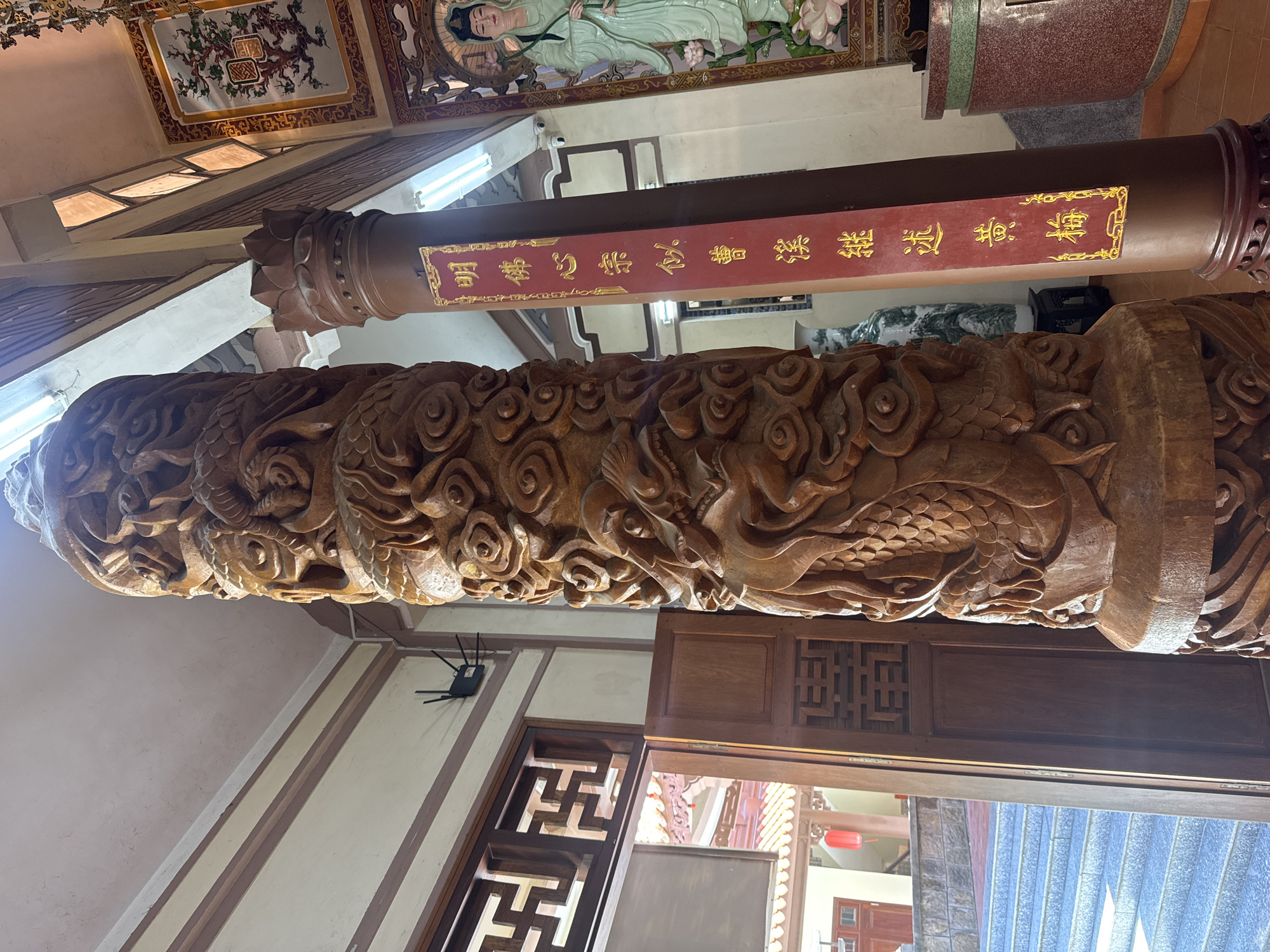 The image shows a detailed wood carving of a dragon on a pillar, likely inside a temple. The carving includes swirling clouds and scales. The pillar is made of dark brown wood and appears aged. In the background, another pillar with a red banner and gold text stands. Above the pillars, a decorative ceiling with a painting of a figure is visible. The setting appears to be a traditional Vietnamese temple. A doorway with ornate woodwork is seen next to the dragon pillar, leading to another part of the temple where an exterior view is visible. The overall location is Nha Trang Ward, Vietnam.