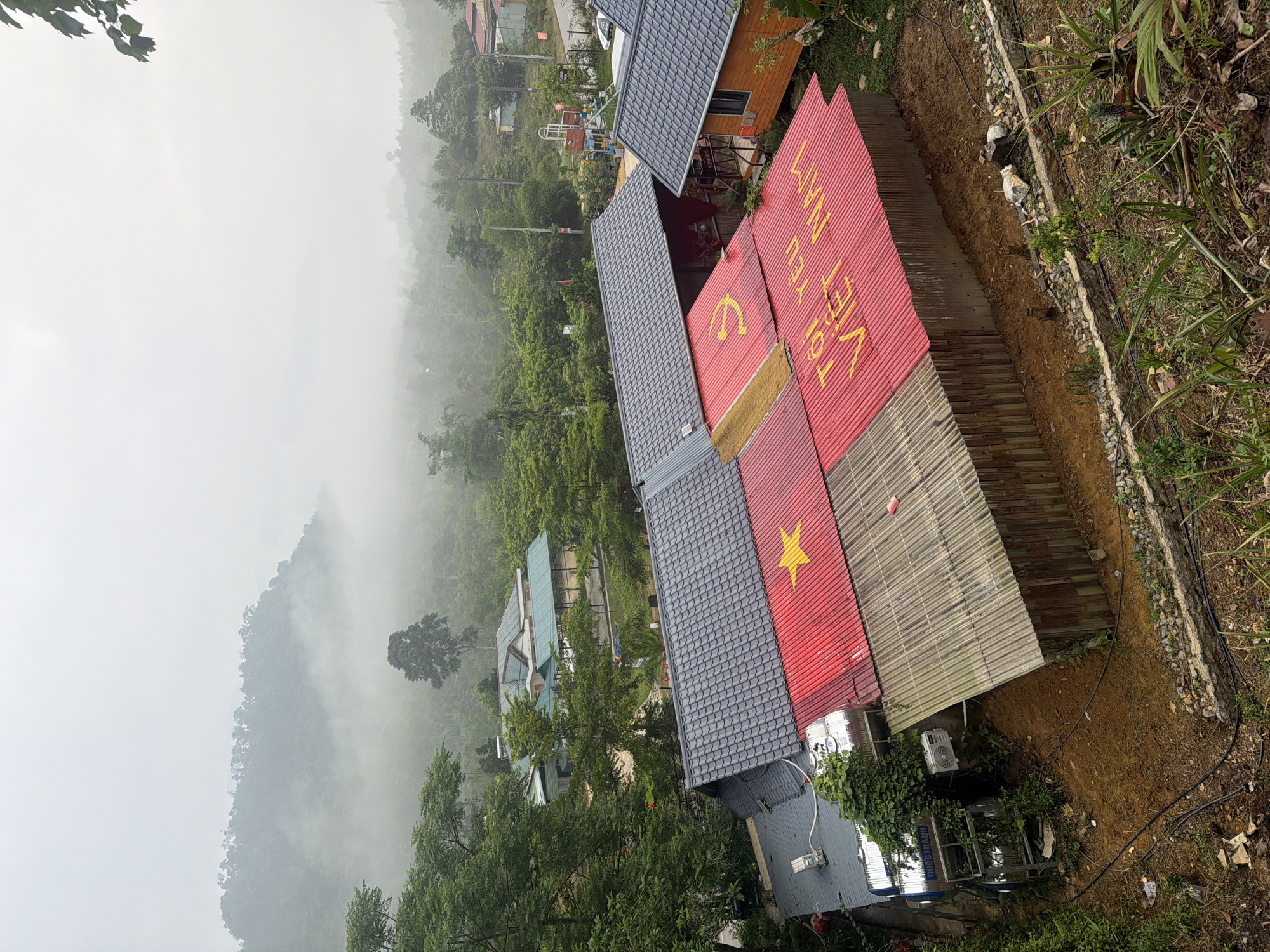 The image shows a hillside scene, likely taken from an elevated position overlooking a misty valley. In the foreground, several buildings with distinct red rooftops are visible, featuring yellow symbols and text in Vietnamese. The symbols appear to be the hammer and sickle, a star, and text that says "Tôi yêu Việt Nam", which translates to "I love Vietnam." The buildings have grey-tiled roofs as well. The background features a lush, green mountainside shrouded in fog or mist, creating a blurred and somewhat ethereal backdrop. Other buildings are visible through the trees. The immediate foreground shows some vegetation and what appears to be a garden area. Location cannot be forensically confirmed as Đà Nẵng; however, the presence of the Vietnamese symbols and architecture indicates a location within Vietnam. No people are visible.