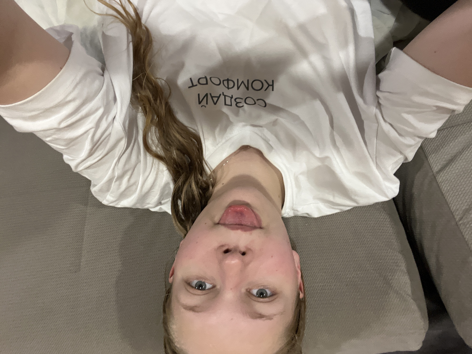 A young woman is lying down on a sofa in a room, looking up at the camera. She has light brown hair that falls around her face and shoulders. She is wearing a white t-shirt with black text printed on it. The text is in Russian and reads "СОЗДАЙ КОМФОРТ", which translates to "Create Comfort". Her facial expression is playful as she sticks her tongue out slightly. The lighting in the room appears to be artificial, suggesting it might be evening or indoors during the day. The background consists of a textured sofa and pillows, common in a domestic setting. The overall mood of the image is casual and lighthearted, captured in a selfie-style shot. The provided location context places this scene in Klepiki, Russia.