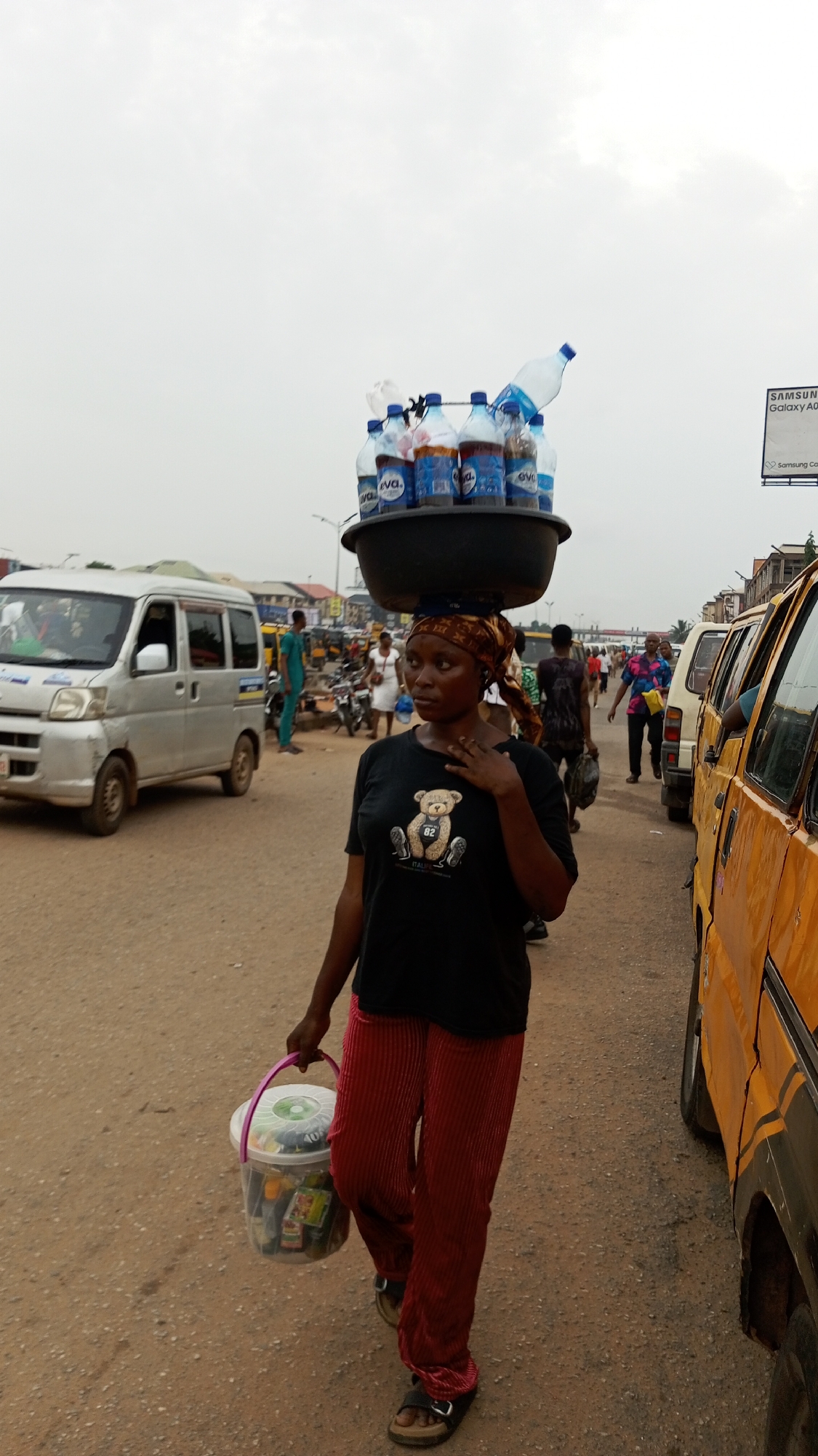 This photograph captures a bustling street scene in Onitsha, Nigeria, centered on a female street hawker. The woman, likely in her late 20s or 30s, walks along an unpaved road with a serious, focused expression. She skillfully balances a large, dark basin filled with numerous plastic bottles, many bearing the "eva." brand label, on her head, secured by a patterned headwrap. One empty bottle precariously rests atop the pile. In her left hand, she carries a clear plastic bucket brimming with various small items, presumably goods for sale.
She is dressed in a black t-shirt featuring a teddy bear graphic, with the numbers "82" and the word "ITALY" visible beneath it. She wears red, vertically pleated trousers and dark, open-toed sandals.
The background depicts a vibrant urban environment under an overcast sky, suggesting either early morning, late afternoon, or a cloudy day. On the left, a silver commercial minivan passes by. To the right, part of an orange/yellow commercial bus is visible. Numerous other pedestrians and motorcycles can be seen further down the road, indicating a busy thoroughfare. In the distance on the right, a billboard partially displays "SAMSUNG Galaxy A0" and "Samsung C.". The scene vividly portrays a common livelihood activity in a Nigerian city.