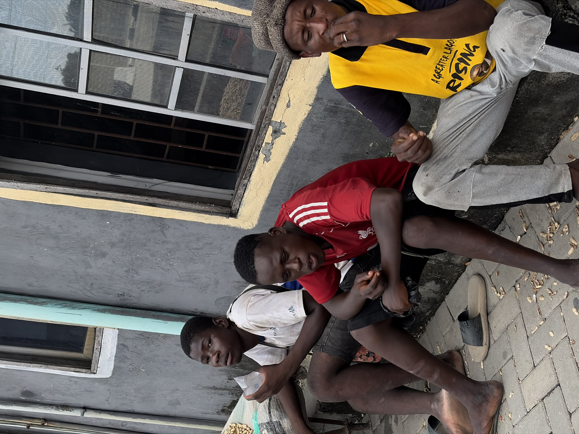In Igboefon, Nigeria, three young men are seated on a raised concrete step adjacent to a weathered building. The building's exterior is primarily grey, with a section painted yellow and a blue-green vertical pipe running along the wall. A window with visible bars behind a screen is present on the building.
The man on the left, wearing a white t-shirt and dark shorts, holds a clear plastic bag containing ice cubes. He is looking slightly upwards and to the left. The middle individual, in a red t-shirt with white stripes on the sleeves and dark shorts, is looking down and to the right, with his mouth slightly open. The man on the right, wearing a dark t-shirt, a bright yellow vest with text reading "A GREATER LAGOS RISING," and light grey pants, has his hand near his mouth and is looking down. He wears a knitted brown cap and a ring on his left hand.
The ground in front of the men is paved with interlocking bricks and littered with discarded peanut shells. A sack filled with peanuts is partially visible on the left. Two pairs of sandals are present on the ground near the men's feet. The overall scene suggests a moment of rest or waiting in an urban outdoor setting.