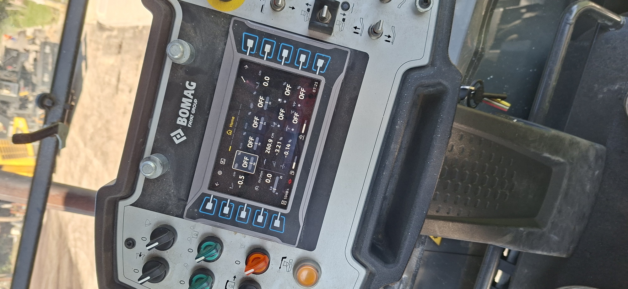 An interior shot of a Bomag Fayat Group compactor's control panel is captured from a slightly elevated, forward-facing perspective. The panel features a digital display screen showing various operational parameters, including "OFF" for multiple functions, speed readings like "0.0 m/min," and measurements in "cm" and "%." Several physical control knobs and buttons are visible surrounding the screen, with two black knobs at the top left, two green toggle switches below them, and an orange rotary dial to the left of the screen. A prominent orange button is located at the bottom left. To the right of the screen, a series of six blue-outlined buttons are aligned vertically. Further to the right, a yellow emergency stop button is partially visible. The control panel is dusty, indicating outdoor use. In the background, out of focus, machinery, possibly other construction equipment, and a dirt or gravel surface are discernible. A set of keys on a keyring hangs from a part of the machine below the control panel. The image was taken in Strazhitsa, Bulgaria.