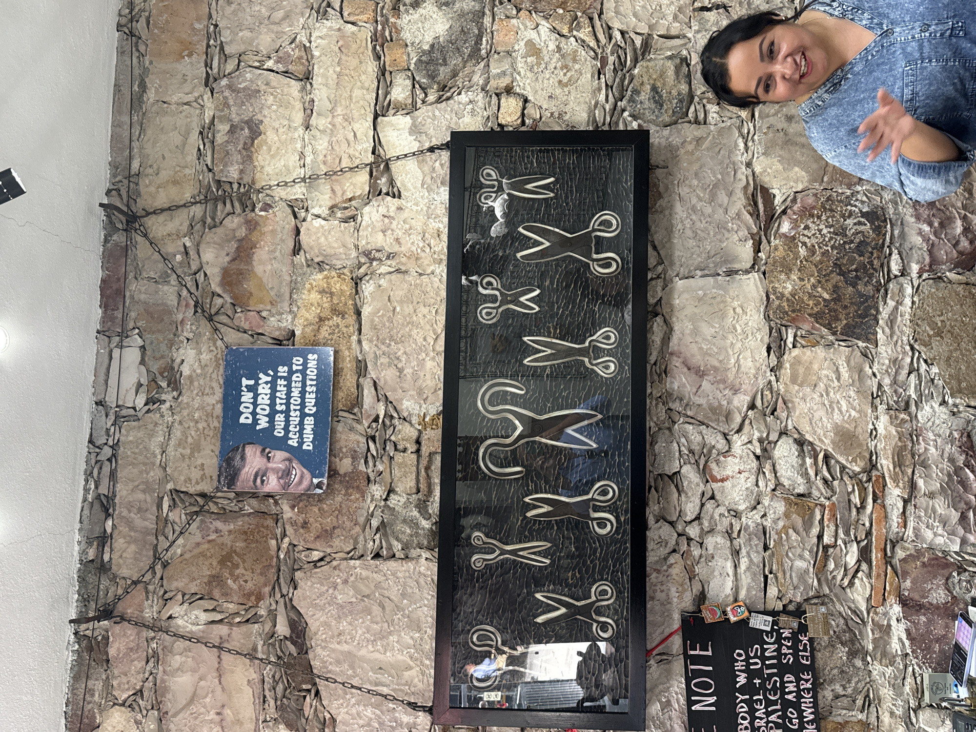The image shows a wall in San Miguel de Allende, Mexico, constructed of roughly hewn stones. Hanging on the wall are several items: a framed collage of scissor images, a sign stating "DON'T WORRY, OUR STAFF IS ACCUSTOMED TO DUMB QUESTIONS" with a picture of a man underneath, and a small black sign titled "NOTE" with handwriting indicating someone who supports Israel and the US should go elsewhere. A woman wearing a denim shirt appears in the upper right corner of the image, smiling and partially obscuring the stonework. The items are suspended by metal chains attached to the wall. The reflection of a person in a blue shirt can be seen at the bottom of the framed collage.