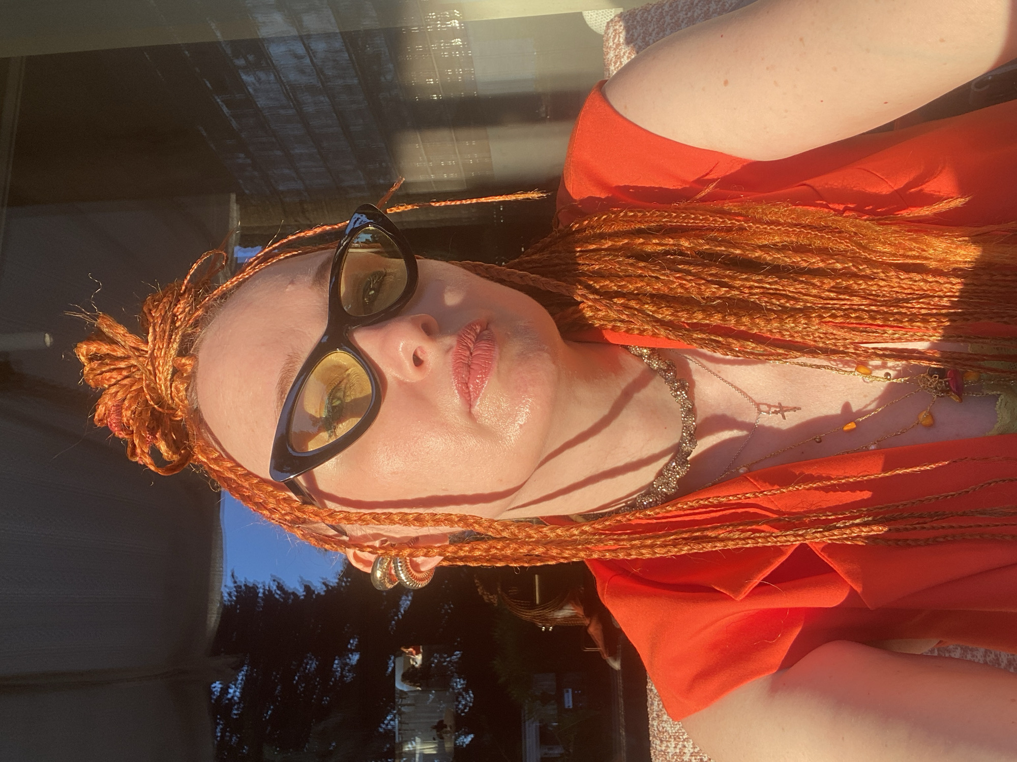 This image depicts an adult female posing for a selfie in the city of Sochi, Russia. She has long, bright orange micro braids, some styled upward on her crown, others hanging down. She wears black cat-eye sunglasses with yellow-tinted lenses, obscuring her eyes. Her lips are full, glossy, and slightly parted. A large, gold-toned double ear cuff adorns her right ear. Around her neck, she wears a dark-toned, textured choker necklace, a thin gold-toned chain with a silver crucifix pendant, and a longer gold-toned chain with irregular orange and amber-colored beads. A fourth, longer chain with a multi-colored pendant is partially visible. She is dressed in a sleeveless, V-neck, bright orange top, possibly a vest or jacket, the fabric appearing structured. She is seated, with a light-colored, textured fabric surface (likely upholstery) visible on either side of her shoulders. Strong, direct sunlight illuminates the scene, creating sharp shadows on her face and body. The background consists primarily of reflections in a large glass window or wall. On the left, reflections show dark, silhouetted coniferous trees and a strip of bright blue sky. On the right, the reflection appears darker with subtle vertical patterns, possibly from an interior or an adjacent building. A faint, indistinct reflection of a human figure or object is visible in the lower left background reflection.