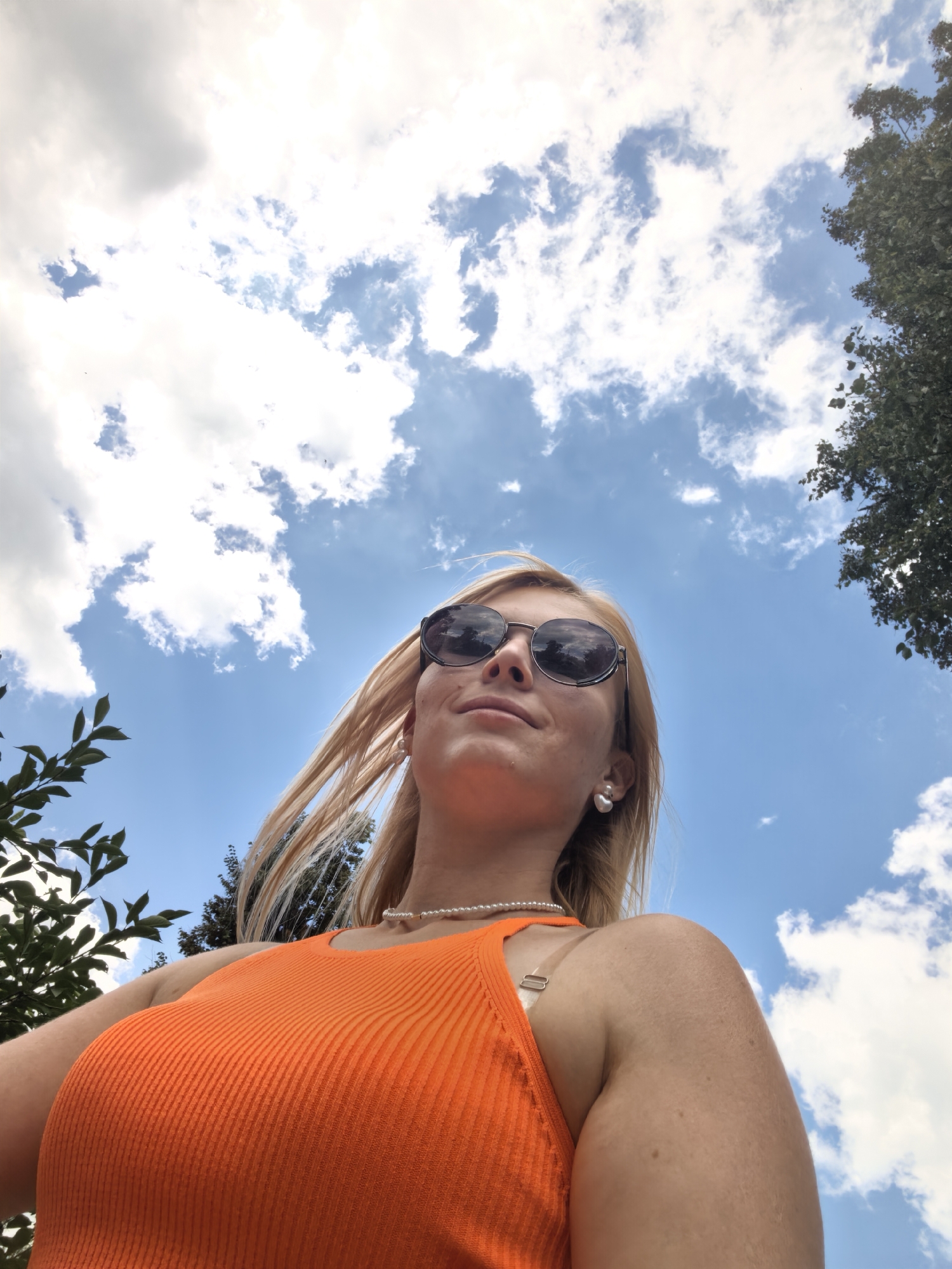 An outdoor, low-angle, wide shot features a blonde woman from the chest up, looking up at the sky. She is wearing large, round sunglasses, a pearl necklace, and an orange, ribbed tank top. Her blonde hair is blowing to the side. The background is a bright blue sky filled with white, fluffy clouds, and the tops of green trees are visible on the left and right edges of the frame. The image was taken in Sterlitamak, Russia.