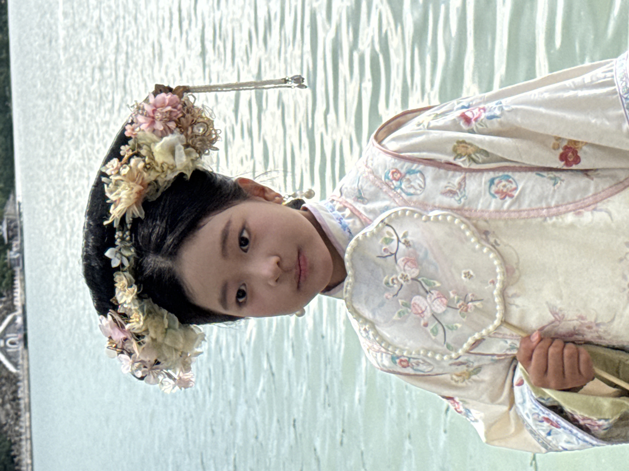 A young East Asian individual, appearing to be a child, is depicted standing outdoors against a backdrop of water and distant land structures. The individual wears a light-colored, long-sleeved garment consistent with historical Chinese attire, featuring intricate embroidery of floral patterns and small figures in shades of pink, blue, green, and red, with pink piping along the seams. A fan-shaped object with a pearl-beaded perimeter and floral embroidery is held in the right hand. The individual's dark hair is styled elaborately with a large, crescent-shaped headpiece adorned with numerous artificial flowers in hues of pink, cream, yellow, and light blue. A beaded chain with small tassels hangs from the right side of the headpiece, and pearl drop earrings are worn. The individual's expression is neutral, with eyes directed towards the viewer. The background consists of a body of light blue-green water with visible ripples. Across the water, in the distance, are trees and several architectural structures, including a prominent white building with a distinct rounded archway. This background environment identifies the location as Kunming Lake within the Summer Palace in Haidian District, China.